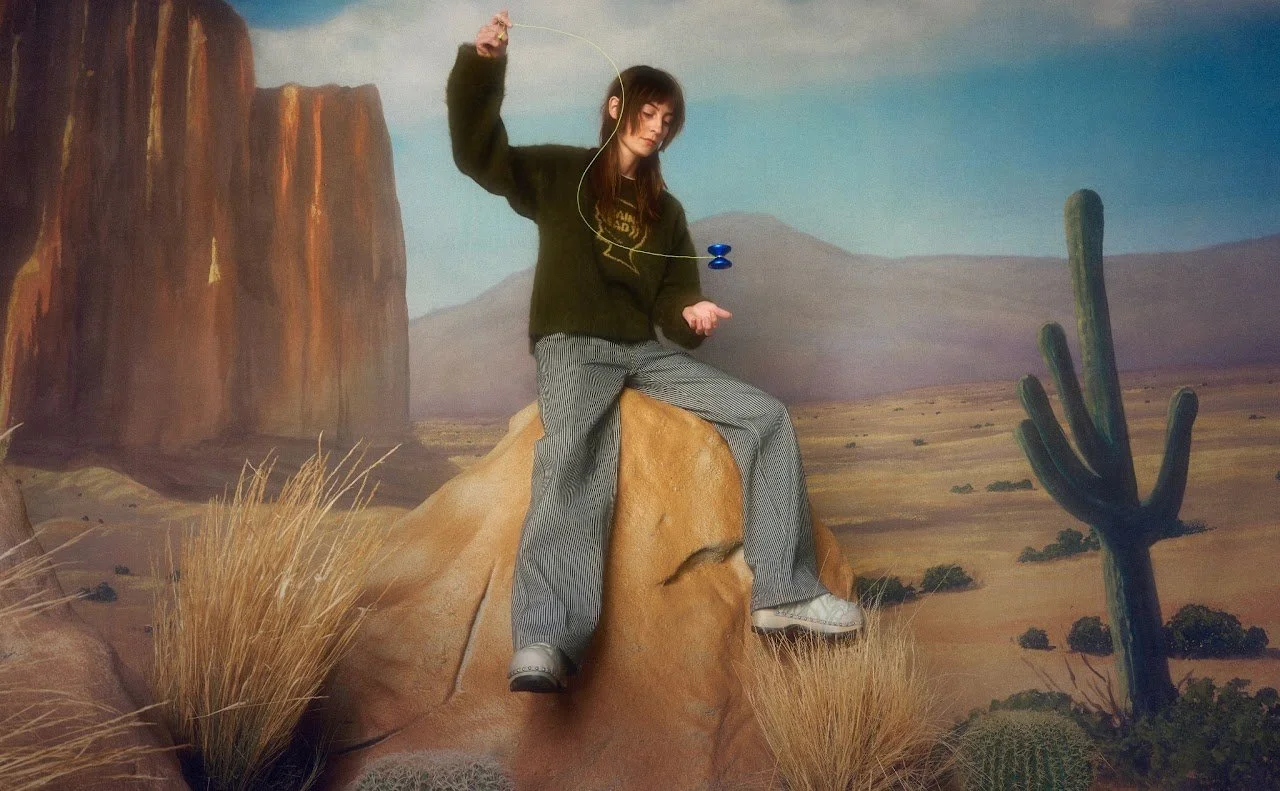 Person sitting on a faux rock in a desert scene, holding a yoyo. Background features cacti, dry grasses, and canyon walls painted scenery.