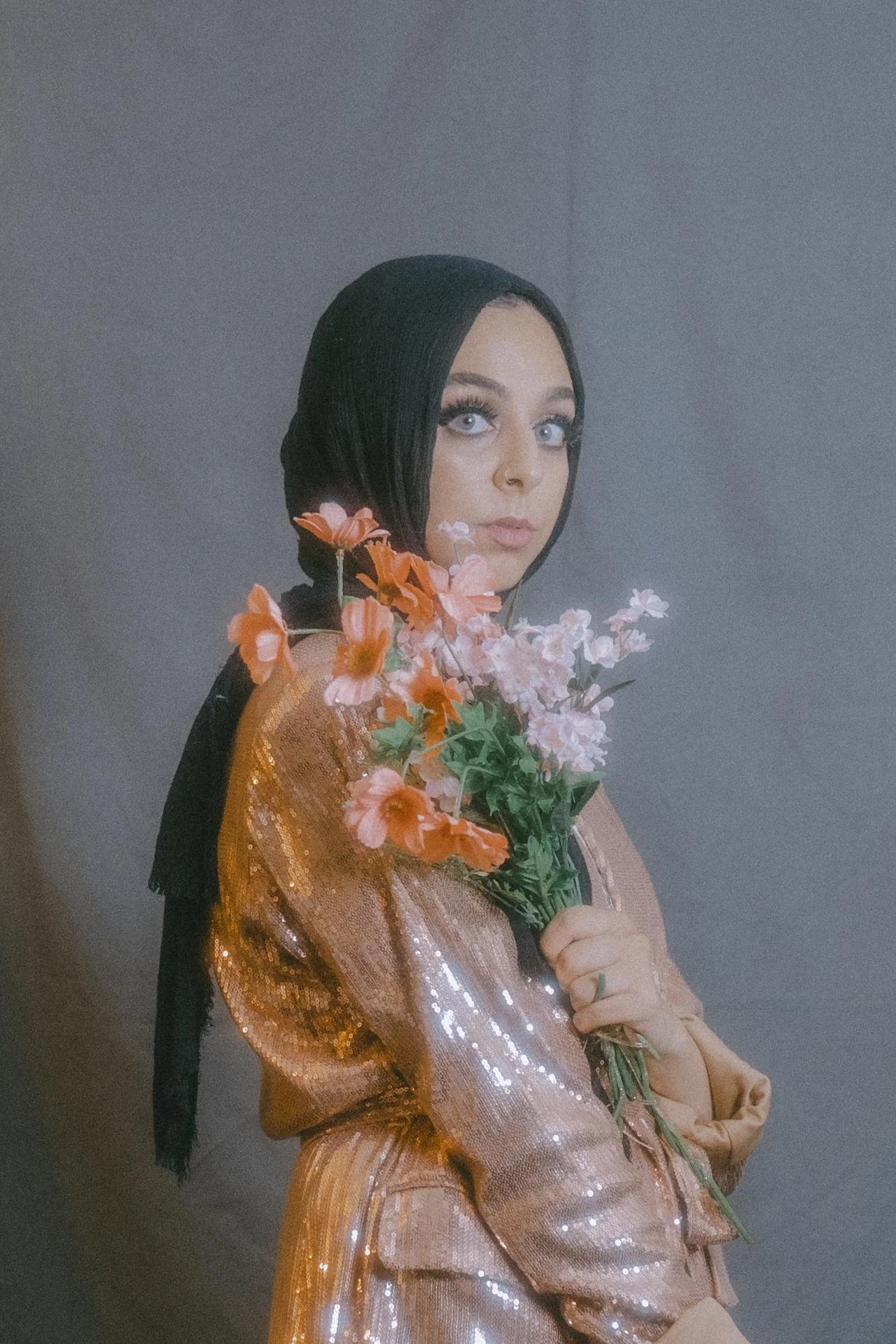 A woman with light skin, black hair, and blue eyes, wearing makeup, holding a bouquet of pink and orange flowers, dressed in a shiny, peach-colored outfit, against a plain gray background.