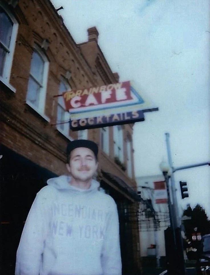 A young man with a mustache wearing a gray hoodie and a black cap standing outside a brick building with signs reading "Gala" and "Cocktails".