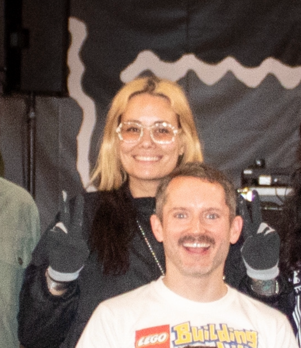 A woman with blonde hair and glasses, smiling, standing behind a man with short brown hair and a mustache, also smiling, both giving a thumbs up.