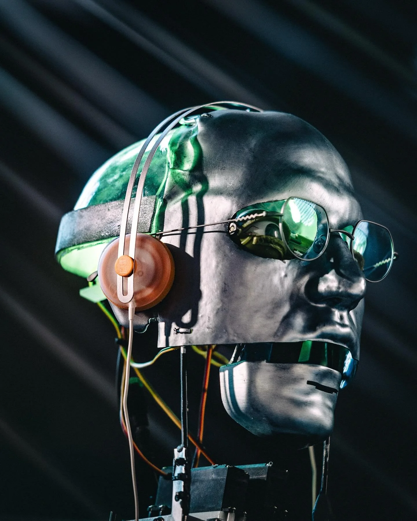 A metallic human head model with electronic components, wires, green-tinted sunglasses, and an orange earphone, set against a dark background with diagonal light rays.