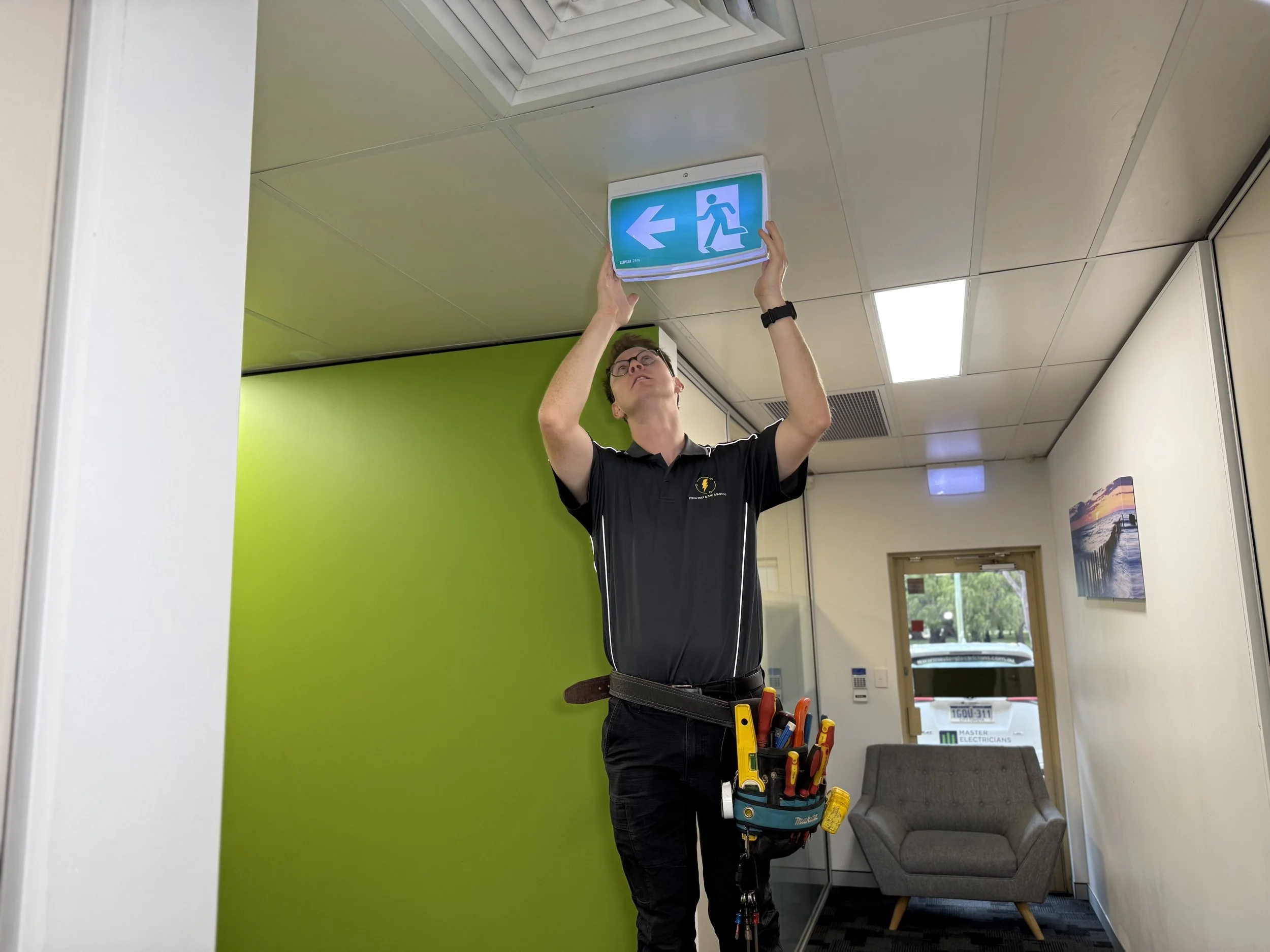 A technician testing completing a 90 minute discharge test of emergency exit signs on the ceiling in a Perth office space.