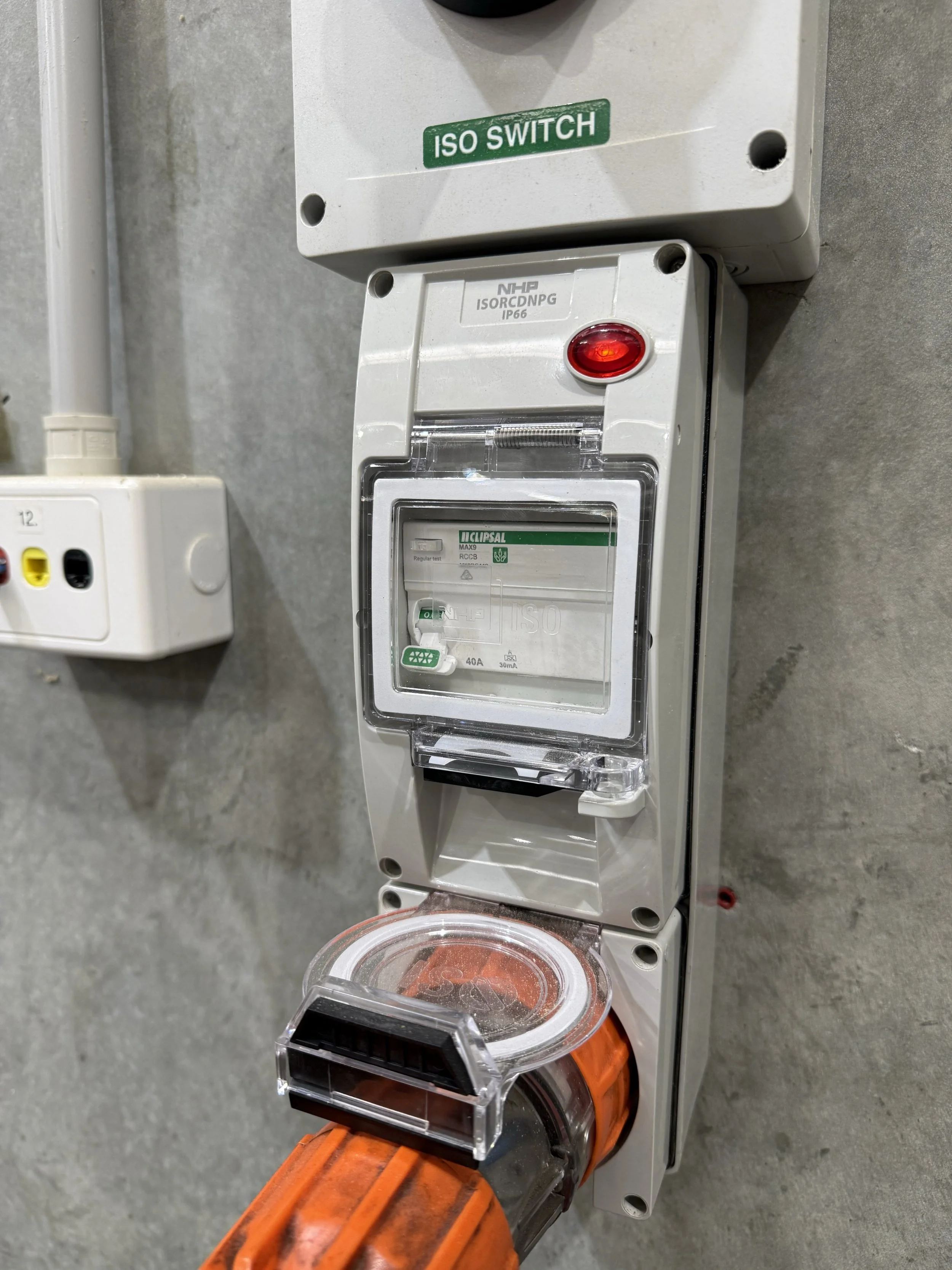 A close-up image of an electrical control box labeled 'ISO SWITCH,' mounted on a wall, with the cover open. A corded power tool with a transparent dust container is shown plugged into an adjacent outlet, with the orange and black power tool partially visible at the bottom of the image.