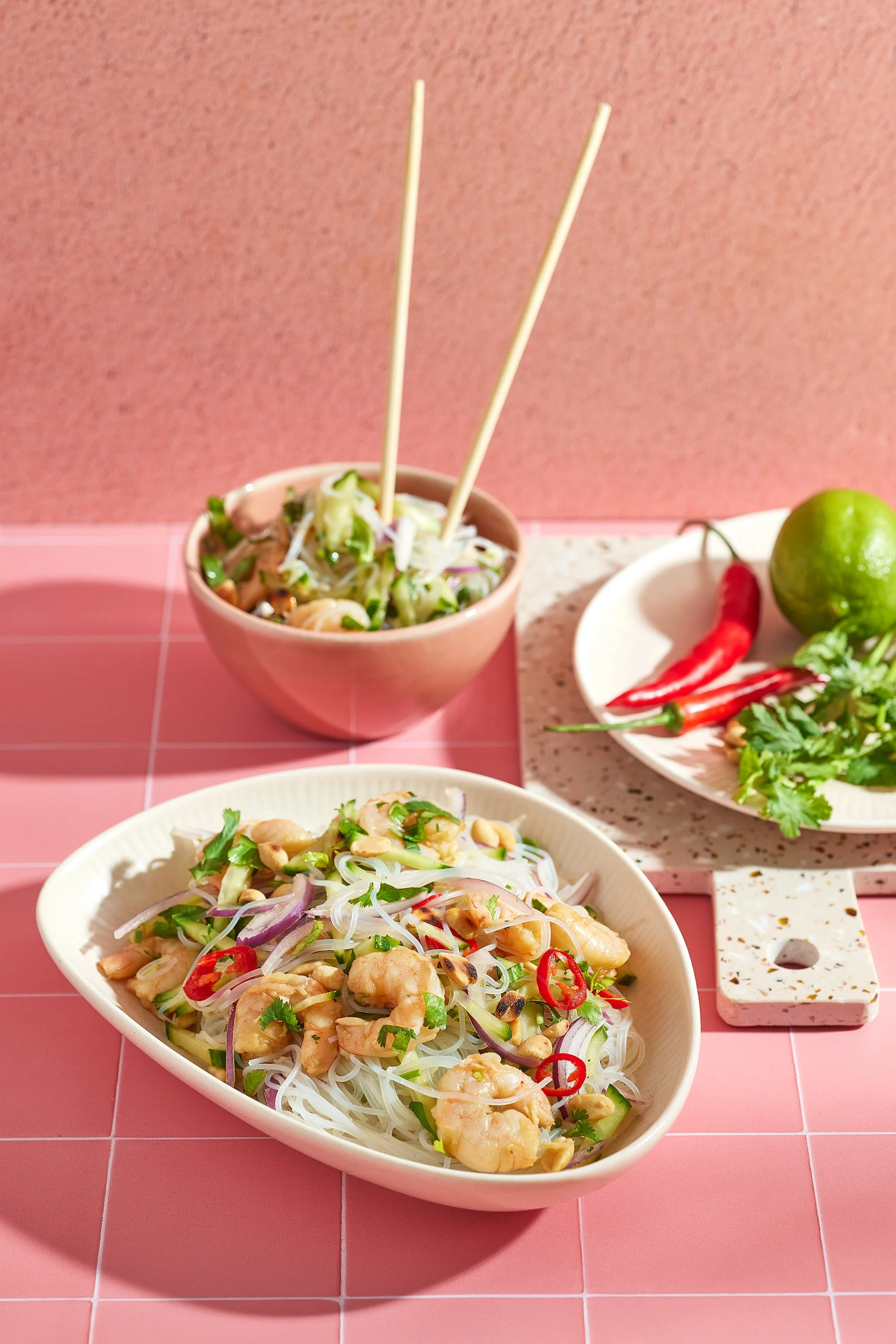 A bowl of noodle salad with chopped vegetables and peanuts. In the background, there are chili peppers, a lime, and cilantro on a white plate, on a pink tiled surface.