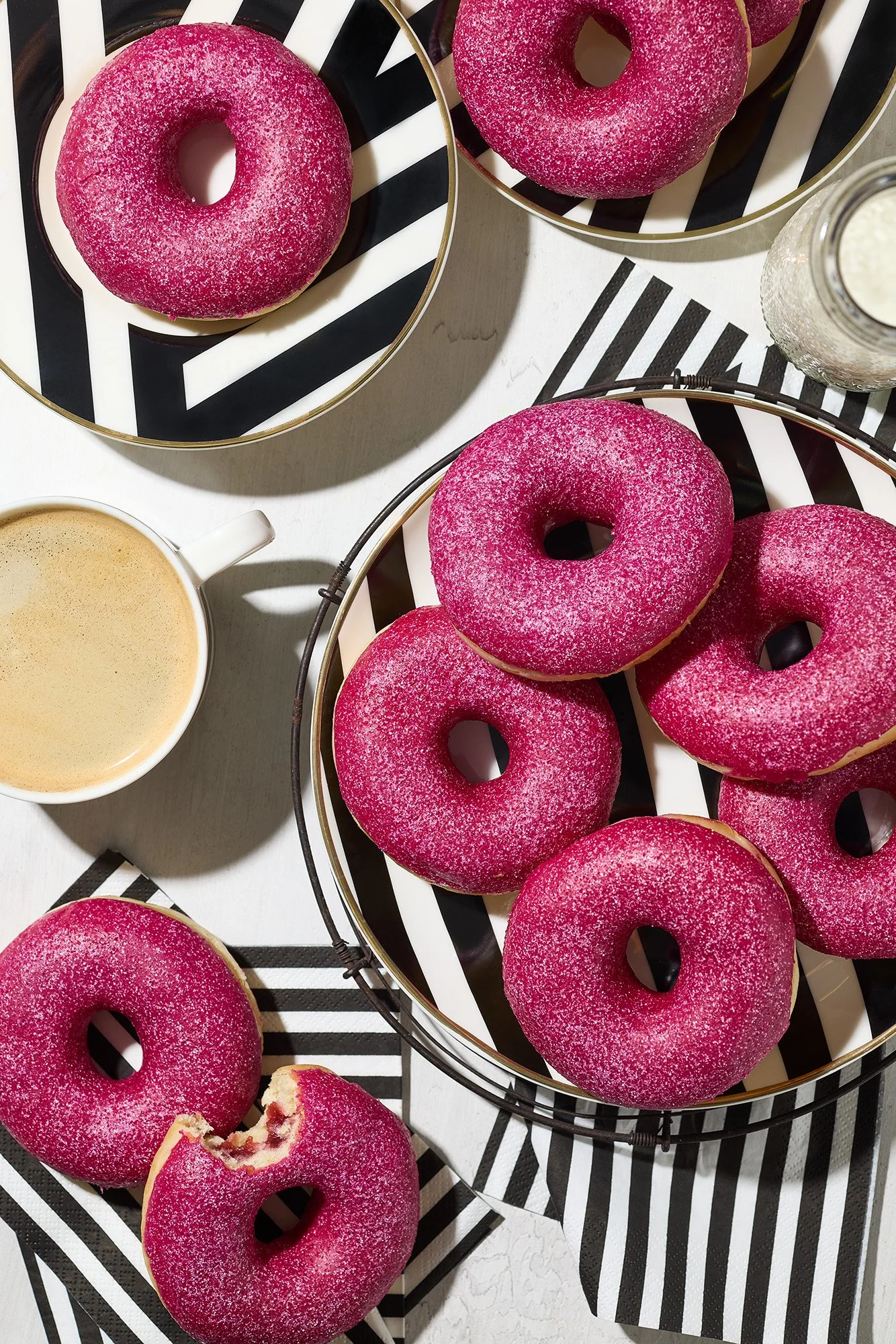 Pink glazed donuts on black and white striped plates and napkins, with a cup of coffee and a jar of milk on a white table.