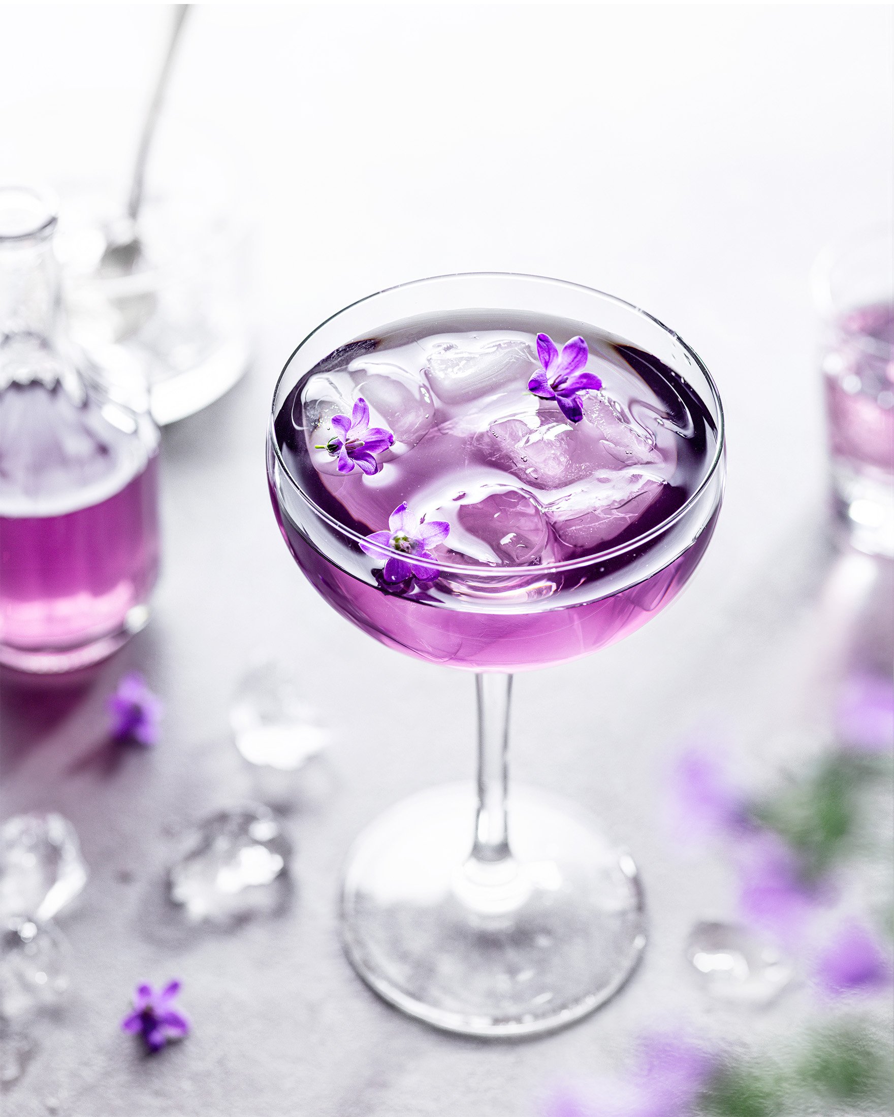 A purple cocktail in a coupe glass with ice and purple flowers, surrounded by scattered ice cubes and purple flowers on a light surface.