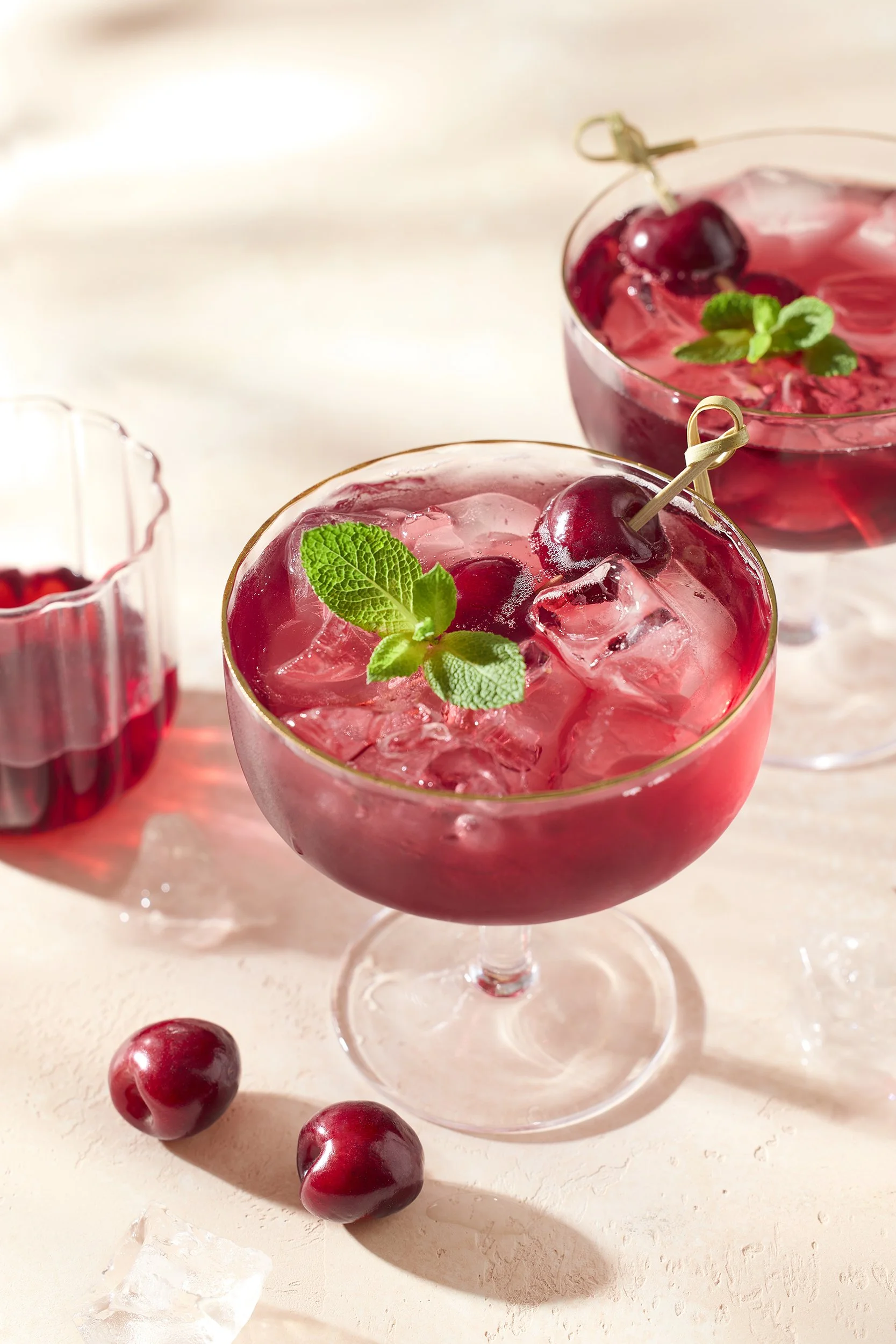 Two glasses of cherry cocktail garnished with cherries and mint leaves, on a light-colored surface with scattered cherries and ice cubes.