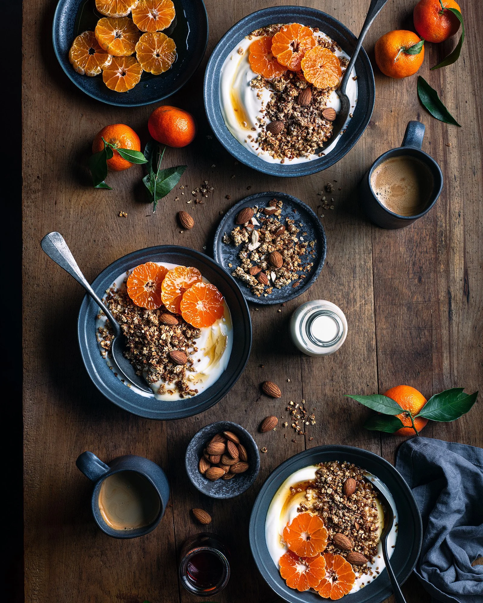 A breakfast scene with three bowls of yogurt topped with granola, almonds, and mandarin orange slices on a wooden table. There are whole mandarins, a small bowl of almonds, a small pitcher of milk, cups of coffee, a small bowl of granola, a small gla