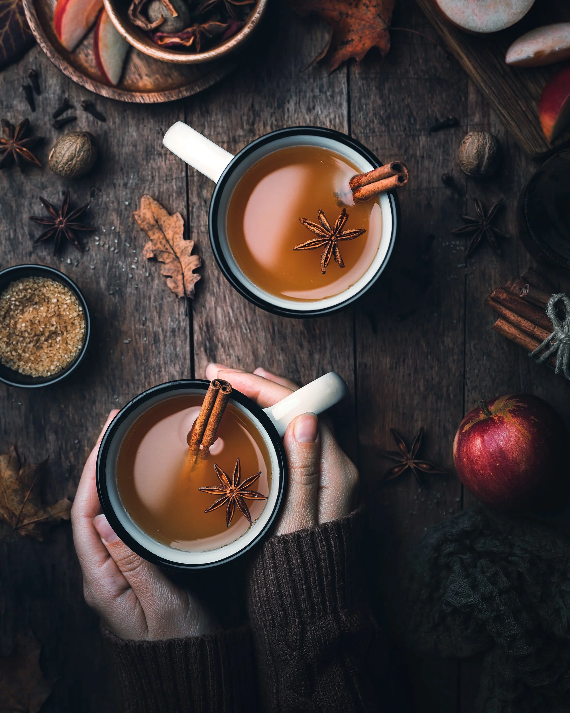 Two hands holding mugs of hot tea or cider with cinnamon sticks and star anise, surrounded by autumn leaves, a red apple, and spices on a wooden table.