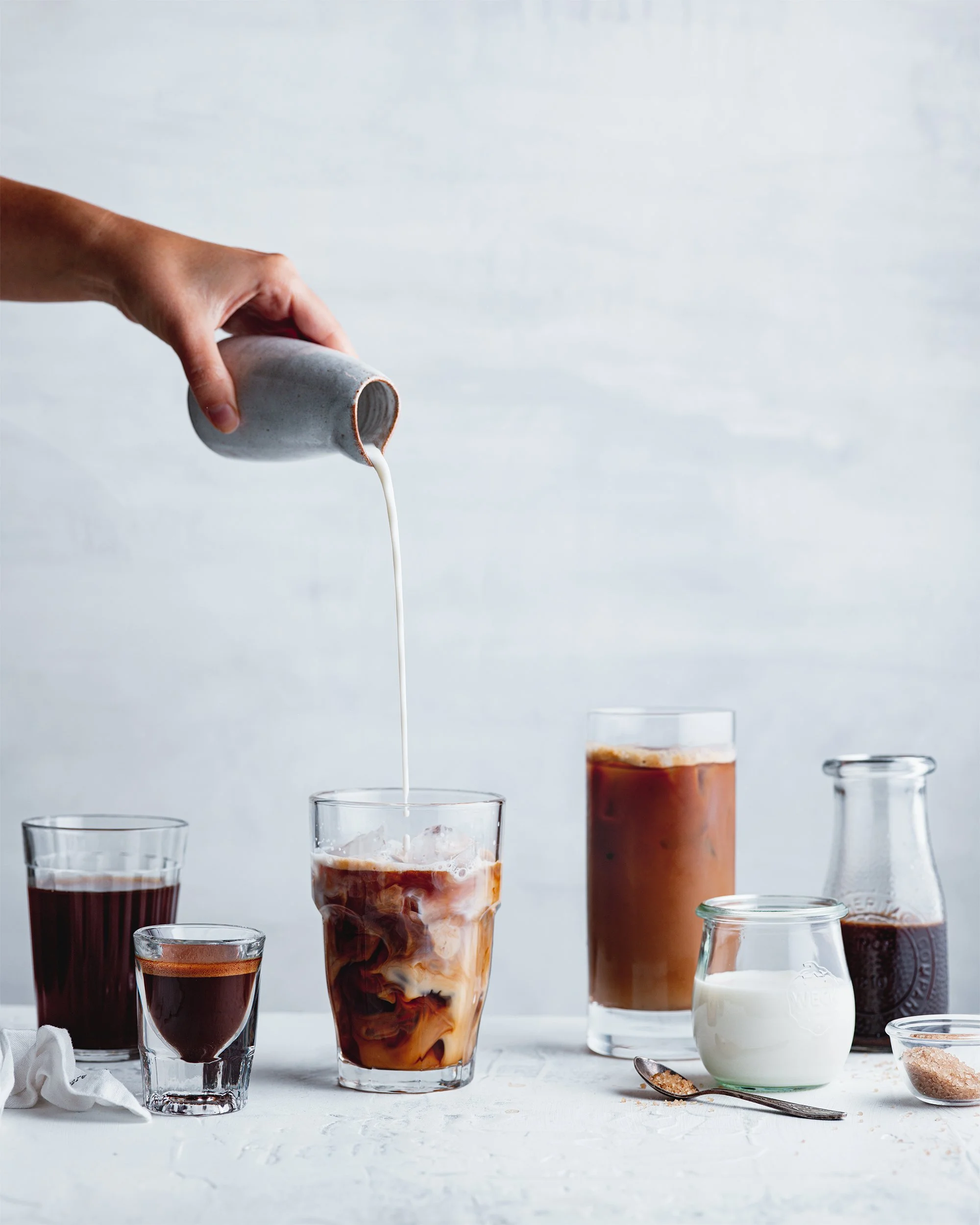 A hand pouring milk into a glass of iced coffee on a table, surrounded by various drinks and ingredients.