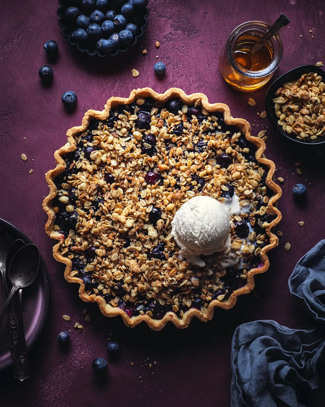 Blueberry pie topped with crushed nuts, served with a scoop of vanilla ice cream, on a dark purple surface with scattered blueberries, honey jar, and a napkin.