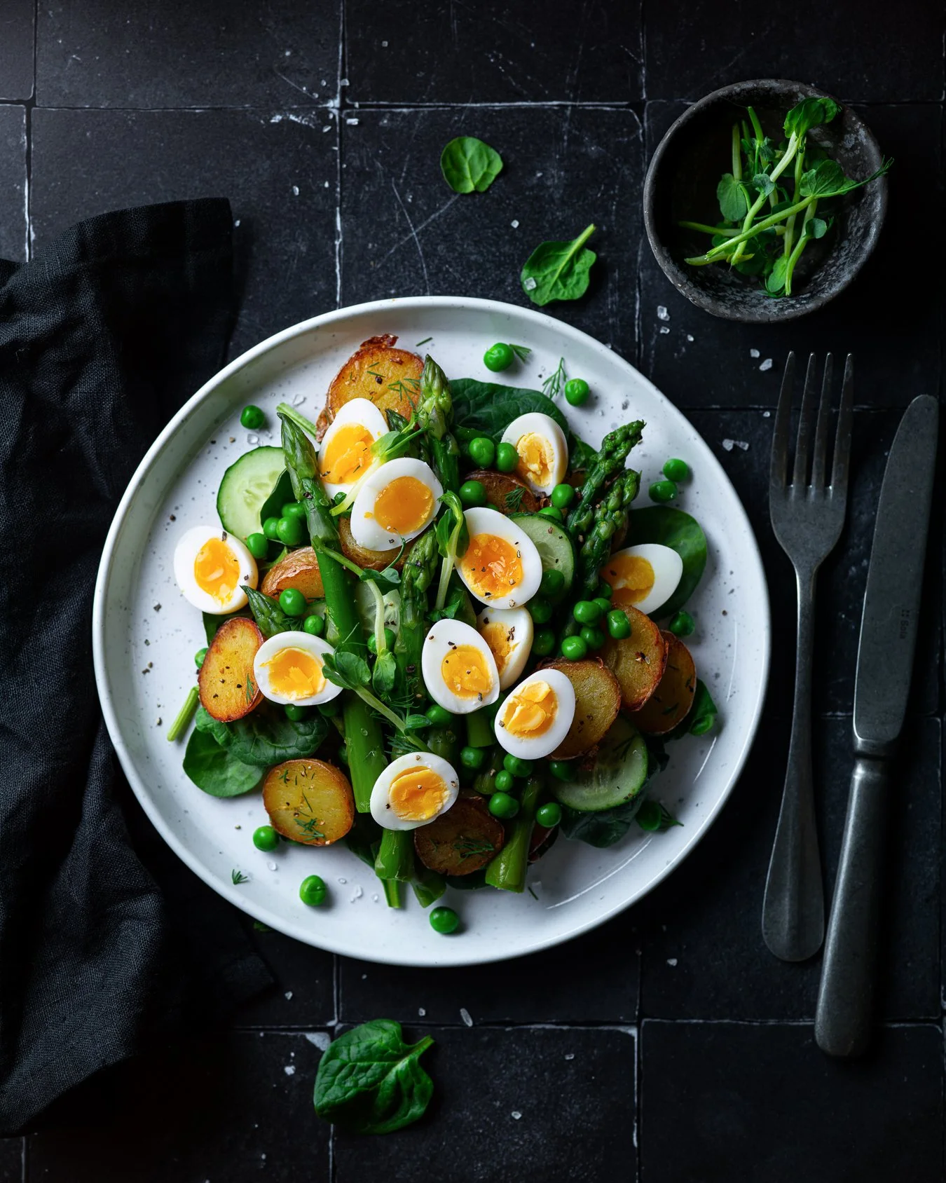 A plate of salad with sliced boiled eggs, green peas, boiled potatoes, cucumber, and spinach leaves, on a black tiled surface with a small bowl of microgreens and a black cloth nearby.