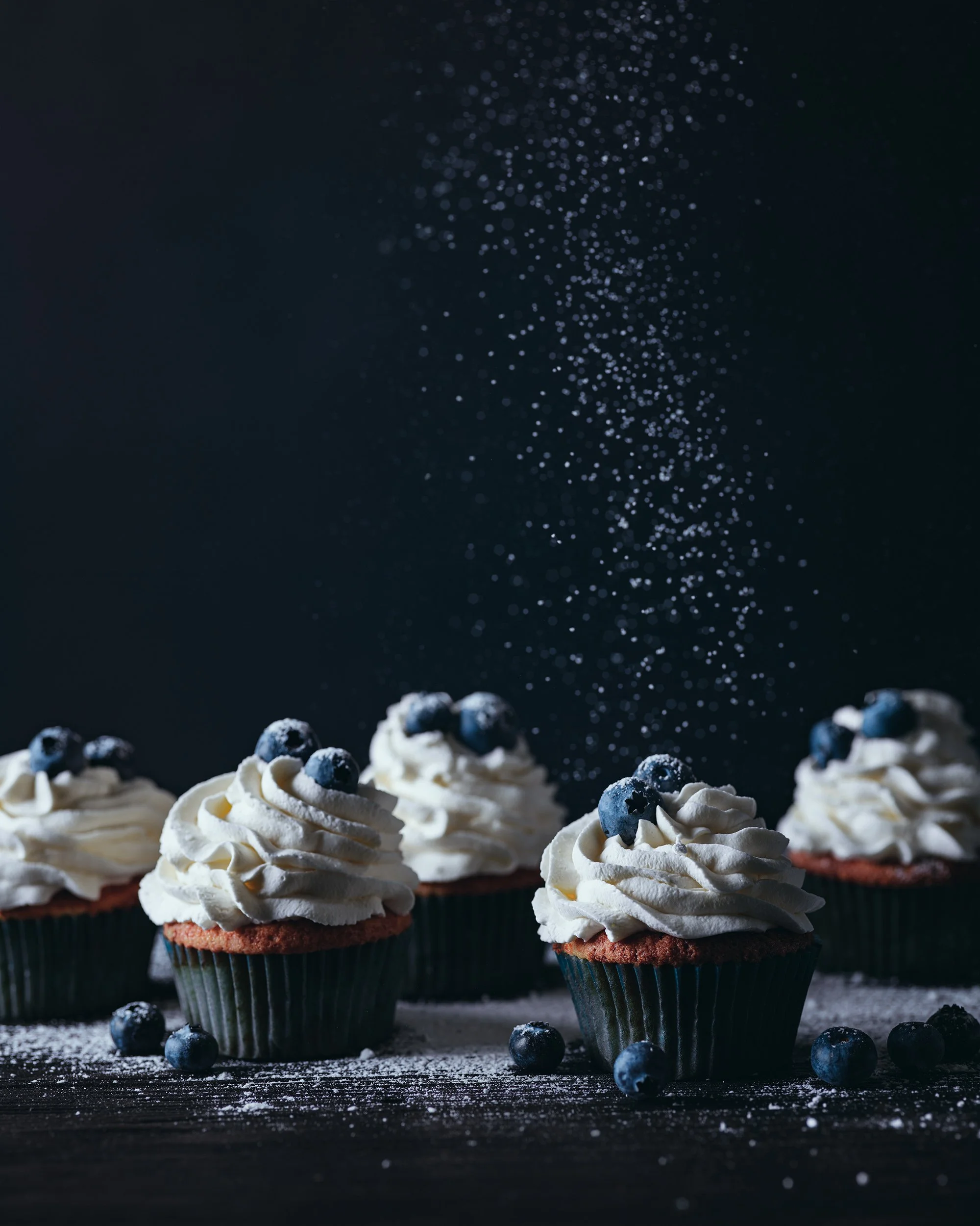 Four cupcakes topped with whipped cream and blueberries, with some blueberries and powdered sugar scattered on the dark wooden surface, and a sprinkle of powdered sugar falling from above.