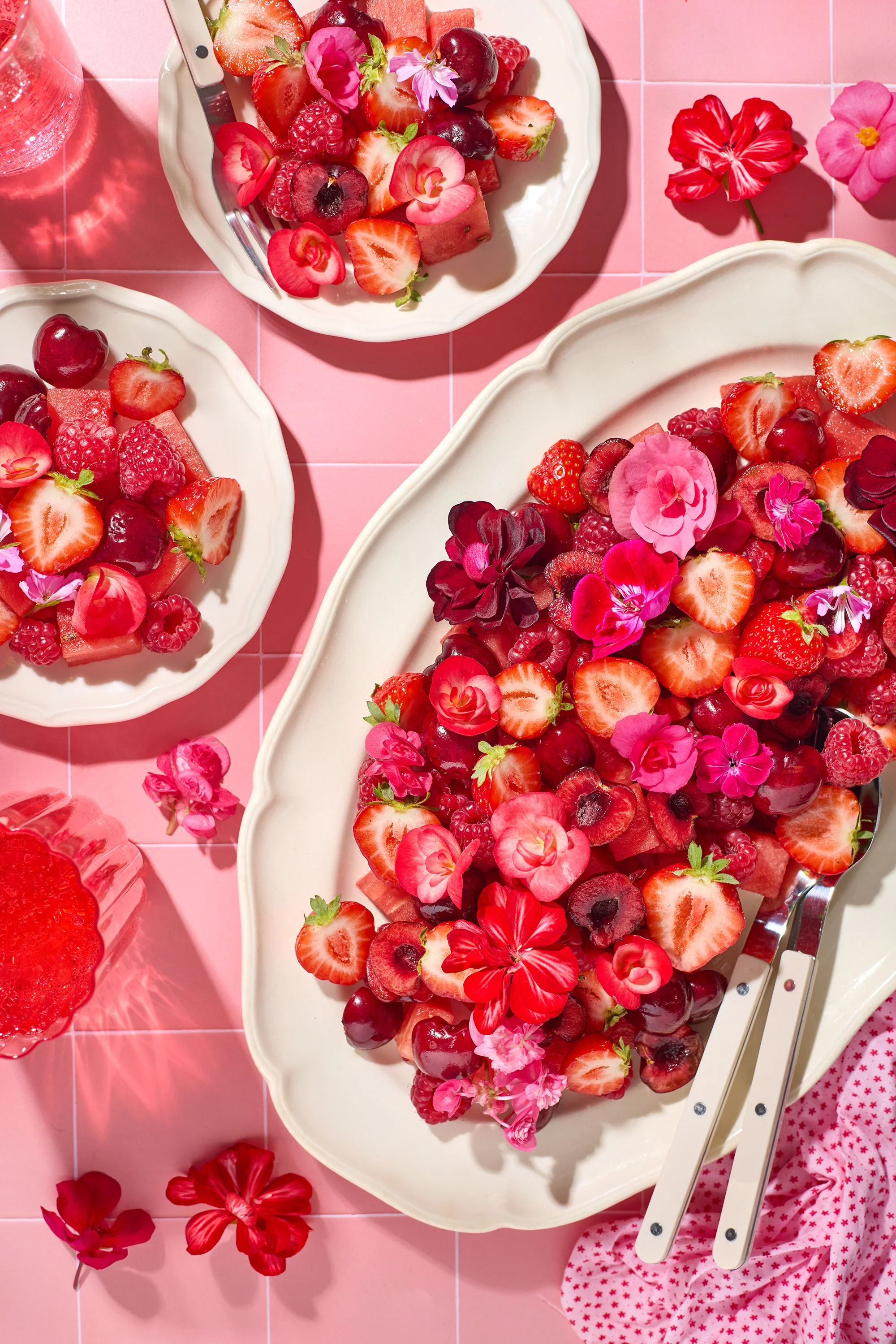 A large white oval platter filled with a colorful array of fresh strawberries, raspberries, cherries, and floral garnishes, set on a pink tiled surface. Smaller white plates with berry arrangements are also visible, along with a pink cloth with red s