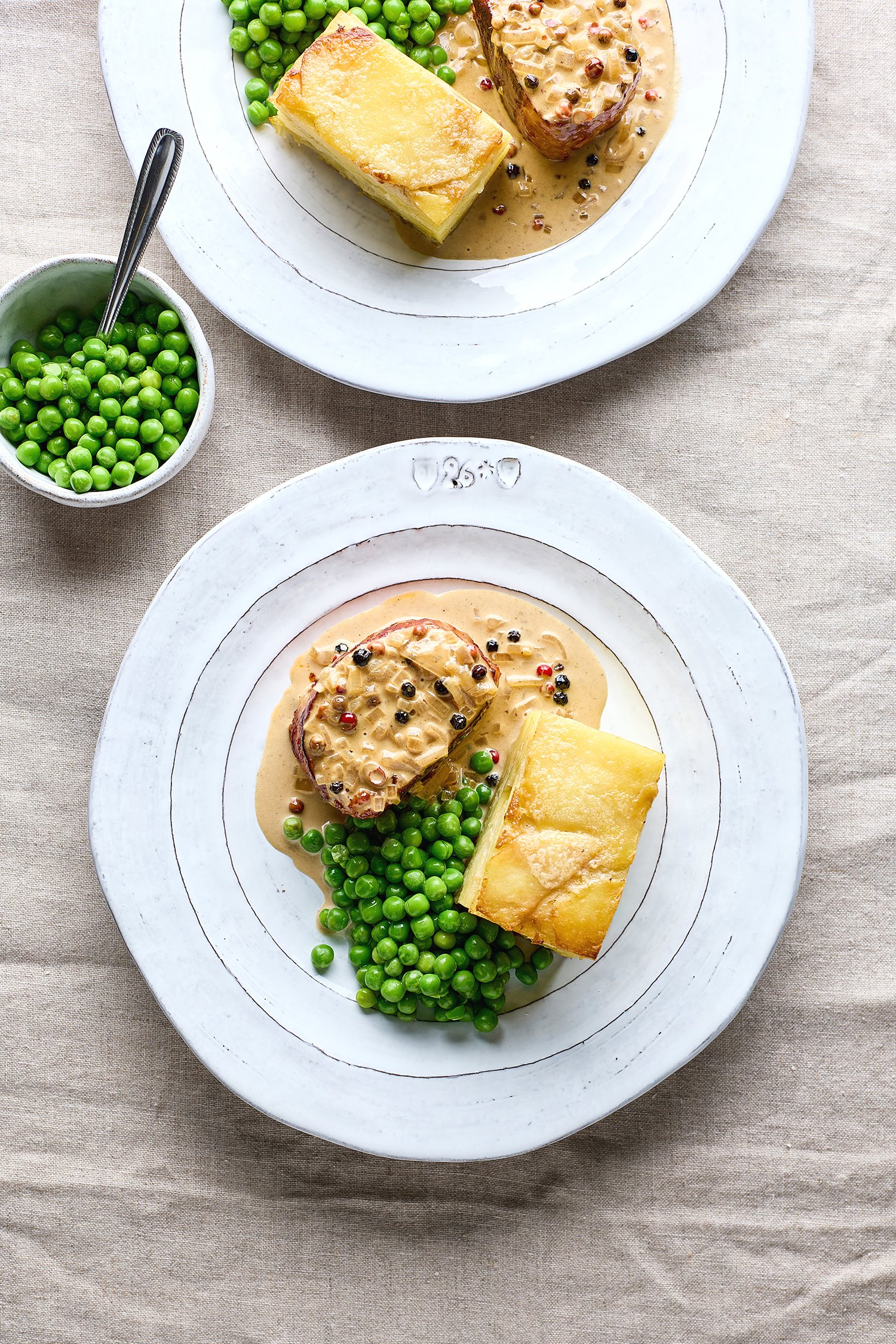 Plate with mashed potatoes topped with beef stew and green peas on a white plate, with additional peas in a small bowl nearby, on a beige tablecloth.