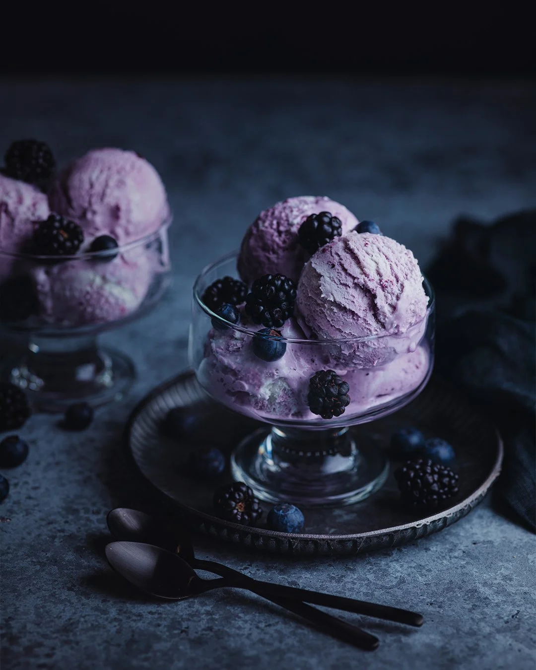 A glass bowl of pink blackberry and blueberry ice cream topped with fresh blackberries and blueberries, placed on a metallic tray with a dark background.