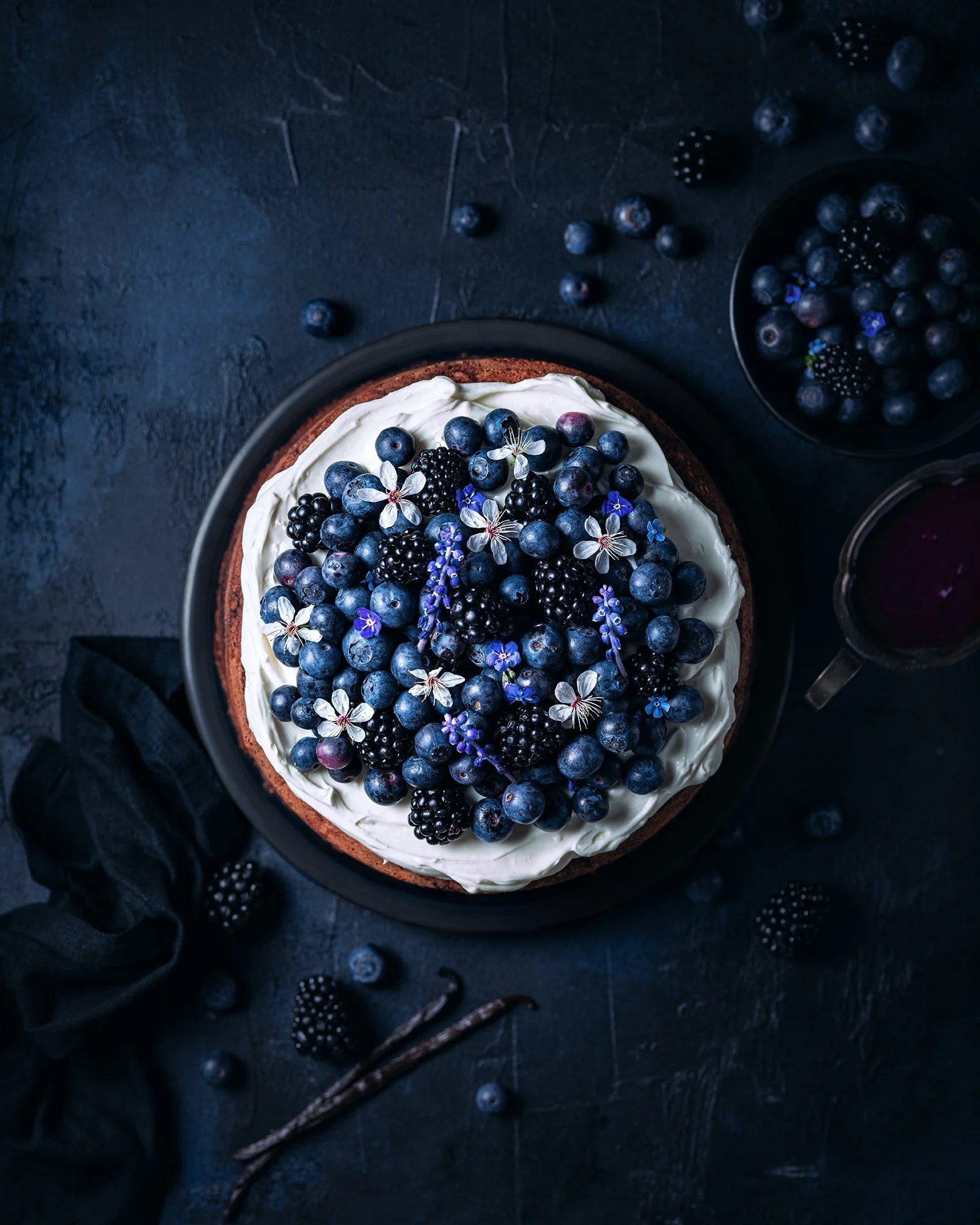 A cake topped with blueberries, blackberries, white flowers, and purple flowers, with additional berries scattered around on a dark surface.