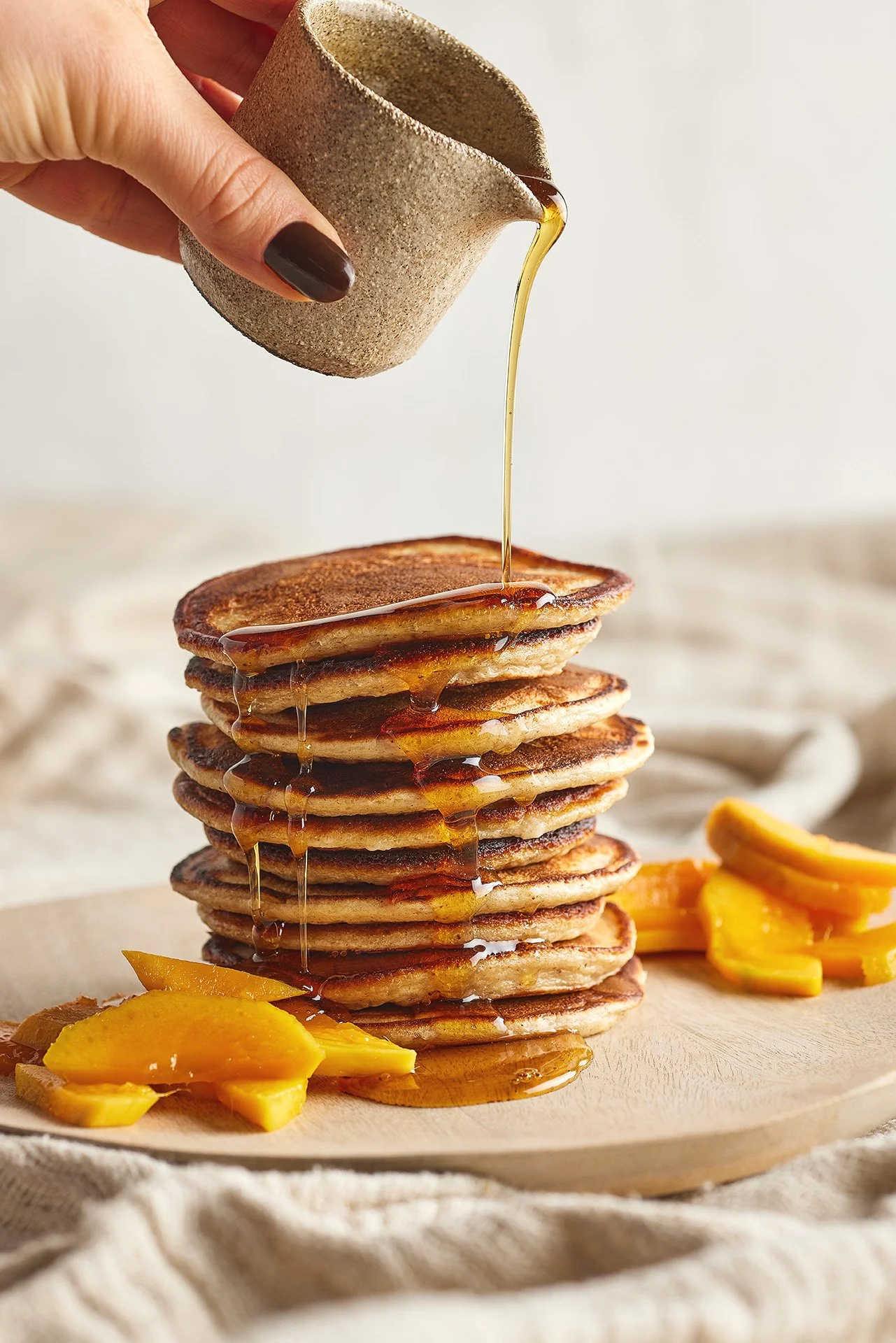 Stack of pancakes with syrup being poured on top, served with sliced mango on the side.