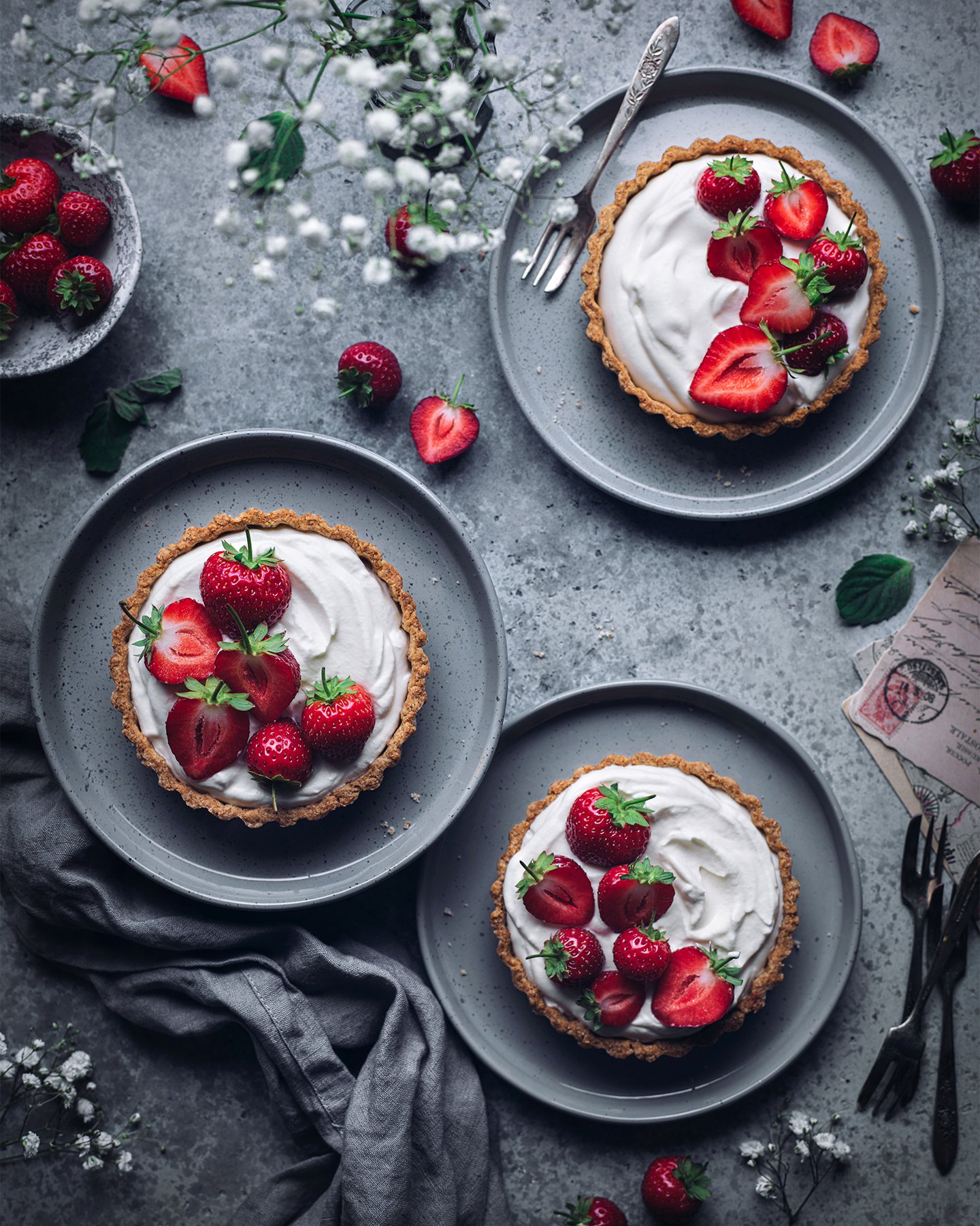 Three strawberry tarts topped with whipped cream and fresh strawberries on gray plates, with additional strawberries and floral decorations on a gray textured surface.