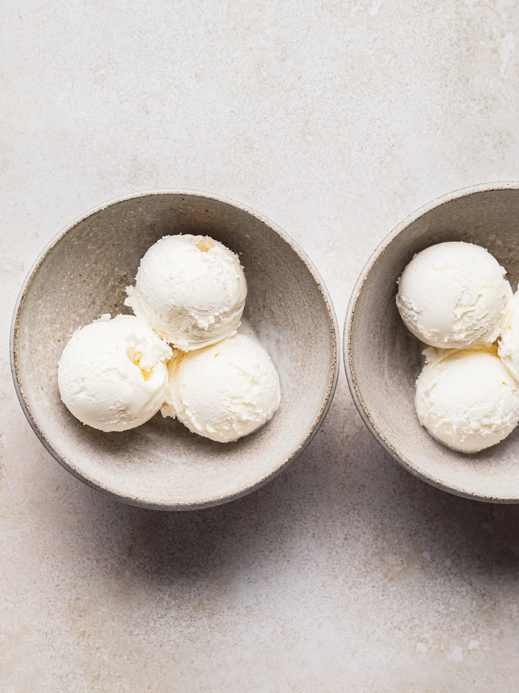 Two bowls each containing three scoops of vanilla ice cream on a light-colored textured surface.