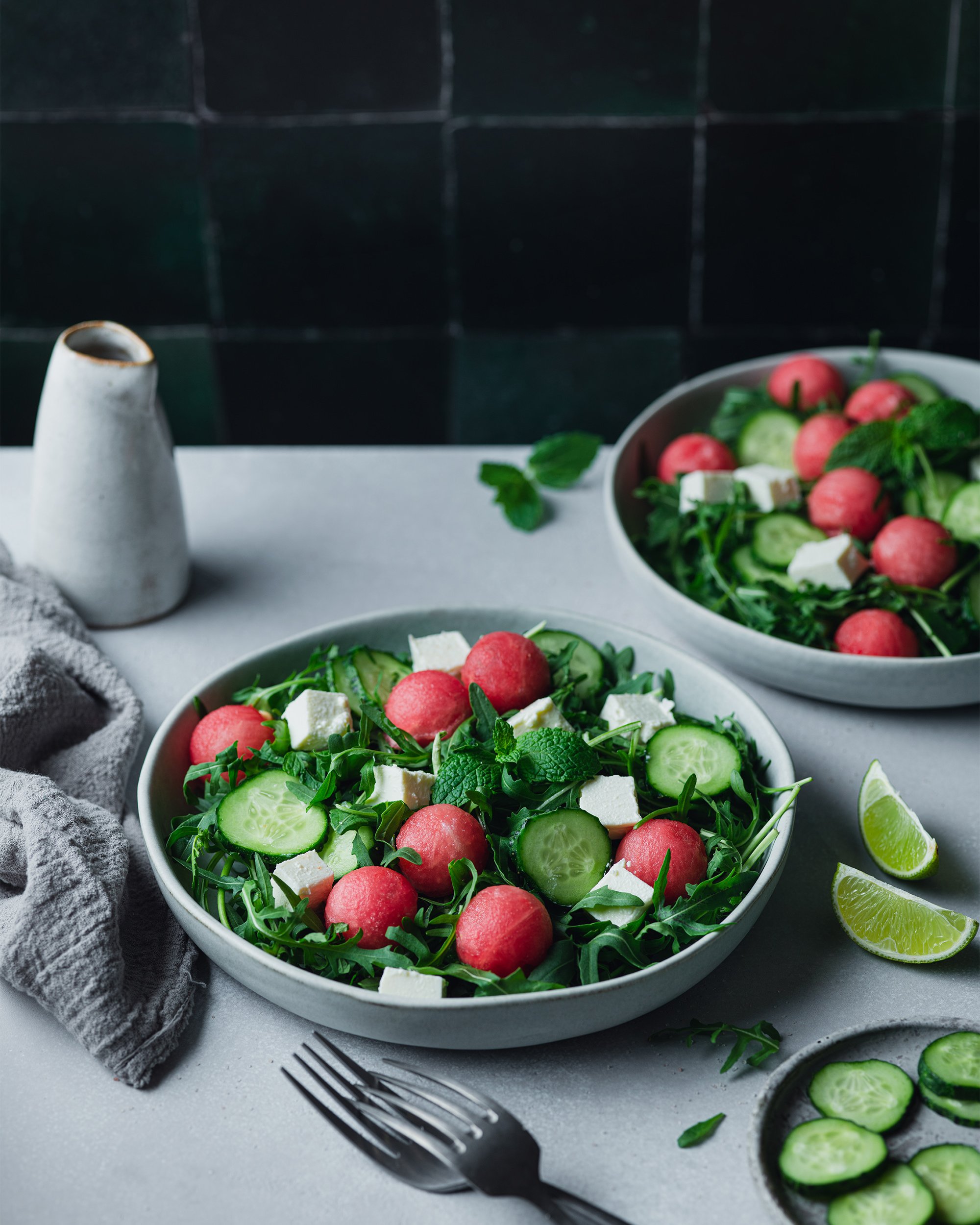 Two bowls of fresh watermelon, cucumber, arugula salad with feta cheese, lime wedges, and sliced cucumbers on a gray table with a fork and gray cloth.