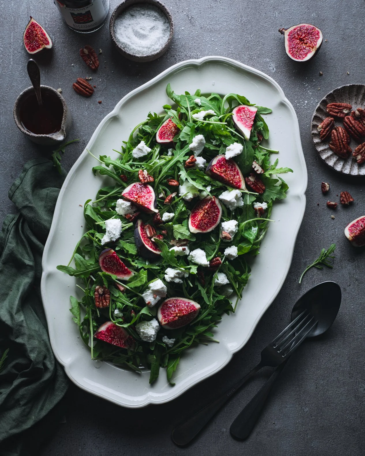 A fresh arugula salad with sliced figs, goat cheese, and toasted pecans on a white platter, surrounded by condiments, a fork, and a green cloth.