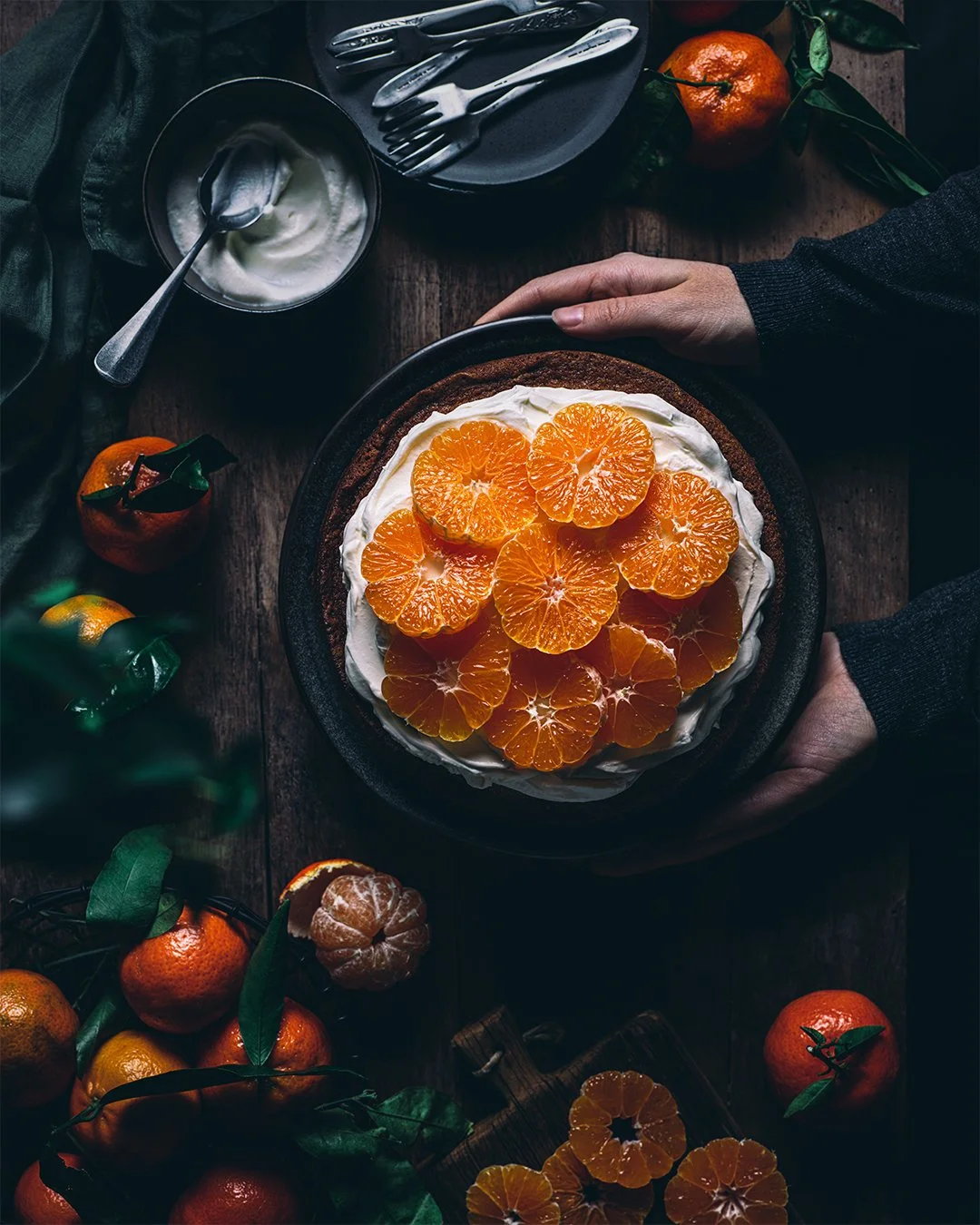A person holding a black bowl with slices of orange on top of a cream or whipped topping. The table has whole tangerines, a peeled tangerine, a bowl of vanilla yogurt with a spoon, and a bunch of tangerines with leaves. There are also utensils and a 