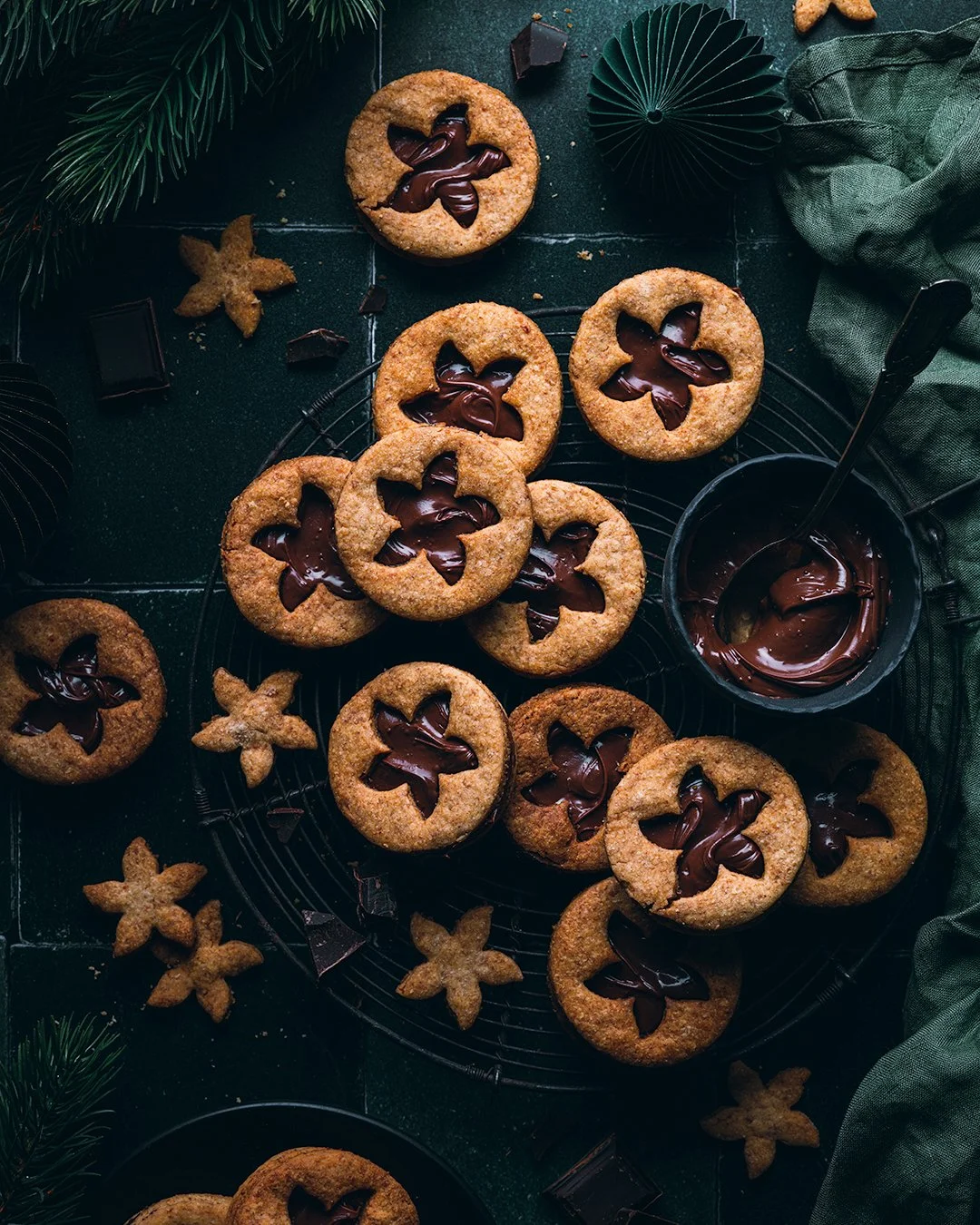 Cookies with chocolate filling on wire cooling rack, surrounded by chocolate squares and holiday decorations.