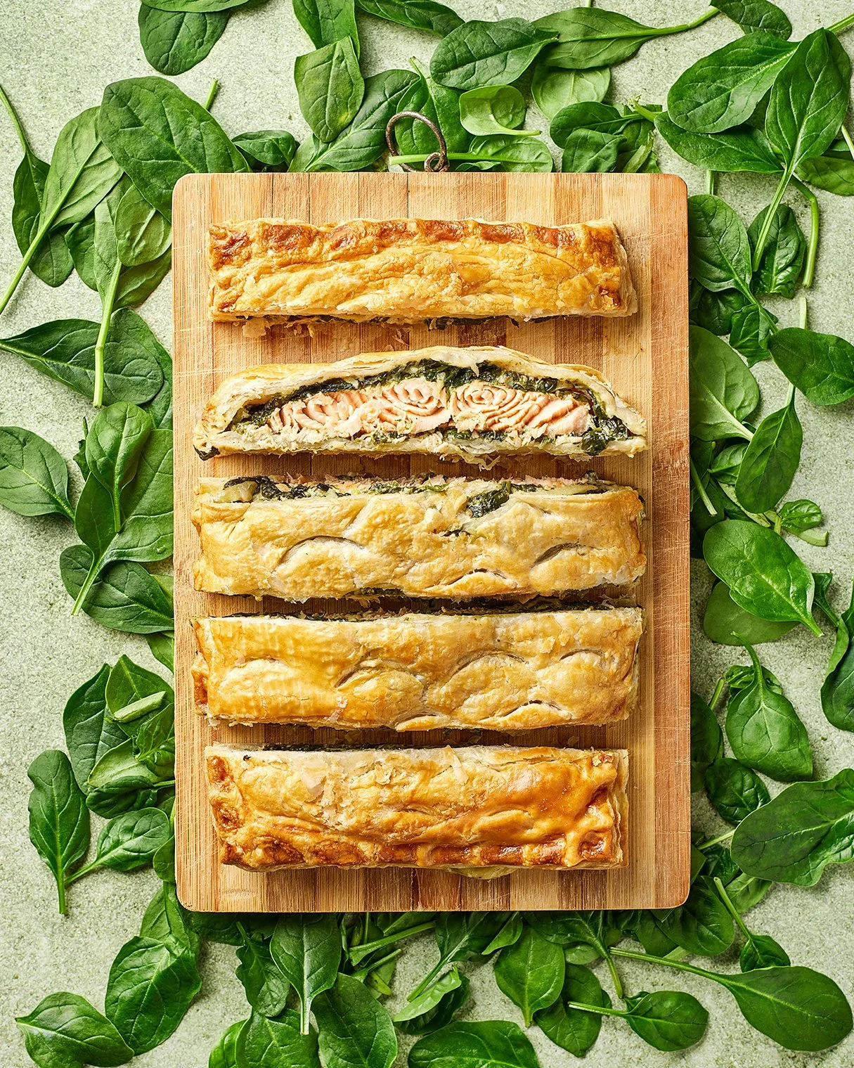 A wooden board with five slices of salmon en croûte, surrounded by green leafy herbs on a light green surface.