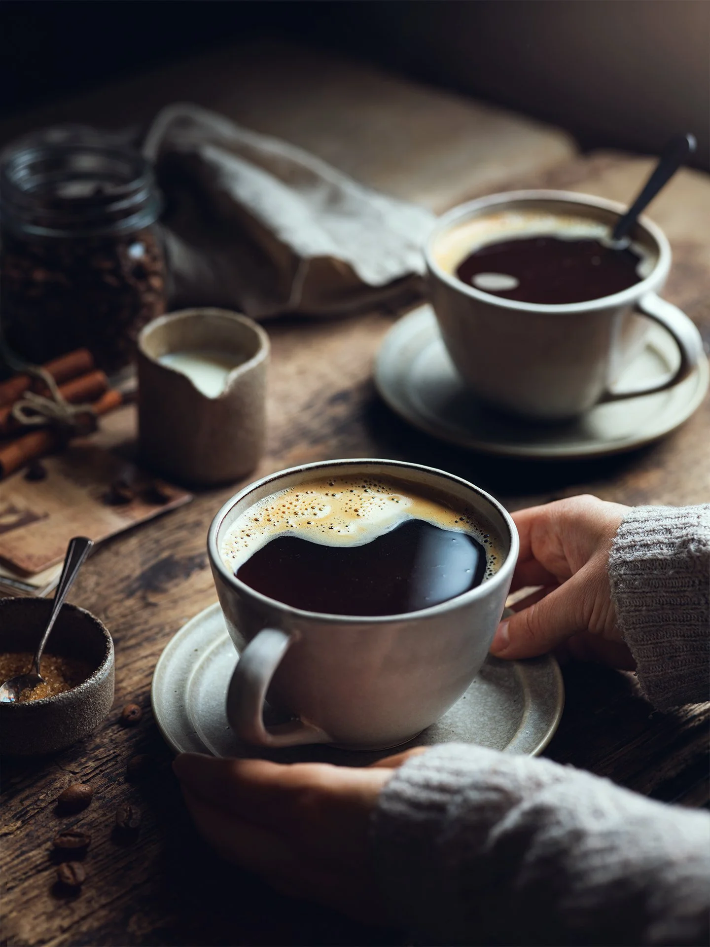 Two cups of black coffee on a rustic wooden table, with a hand holding one cup and various coffee-related items like a jar of coffee beans, cinnamon sticks, and sugar nearby.