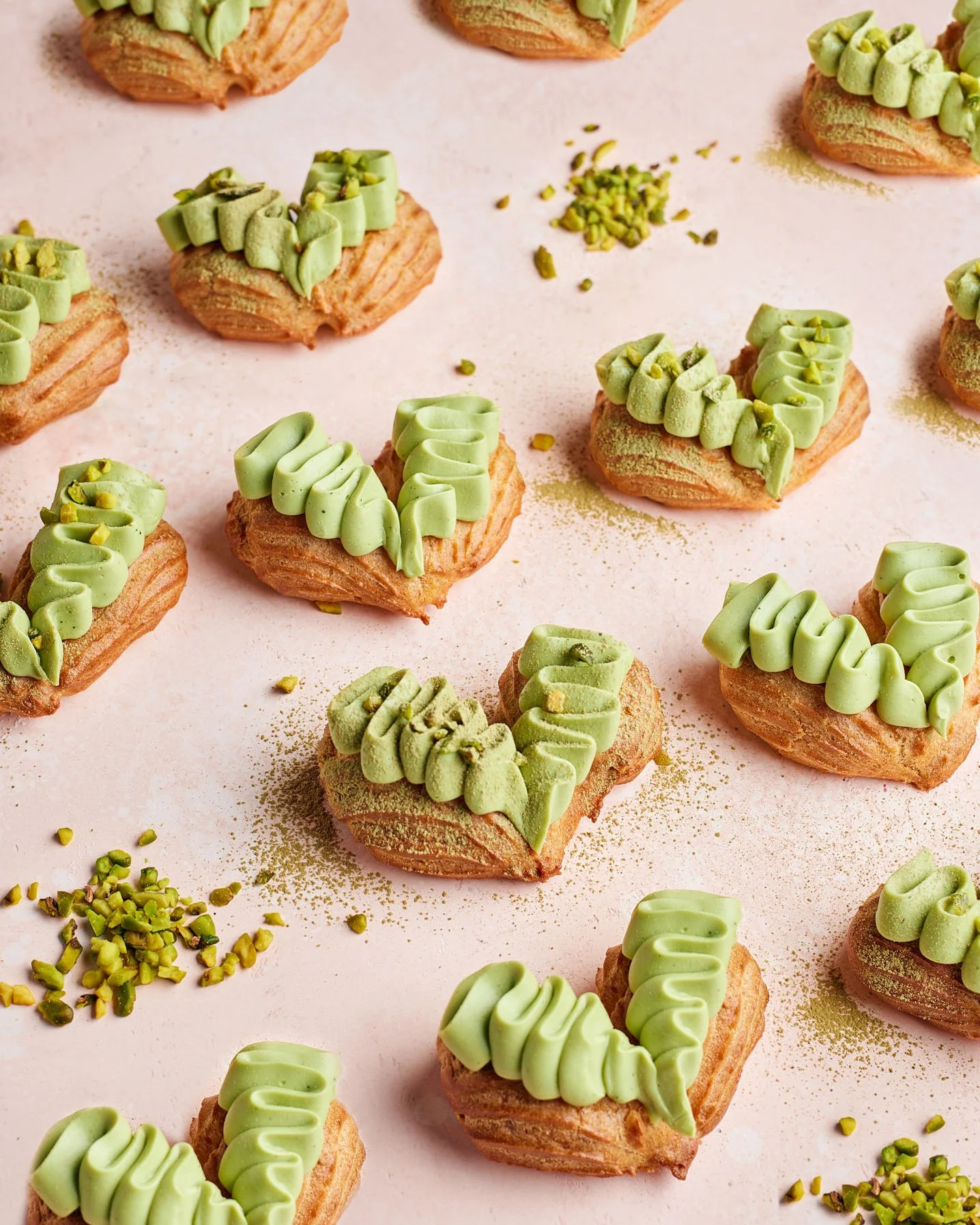 Heart-shaped pastries with light green frosting, garnished with chopped pistachios, on a pink background.