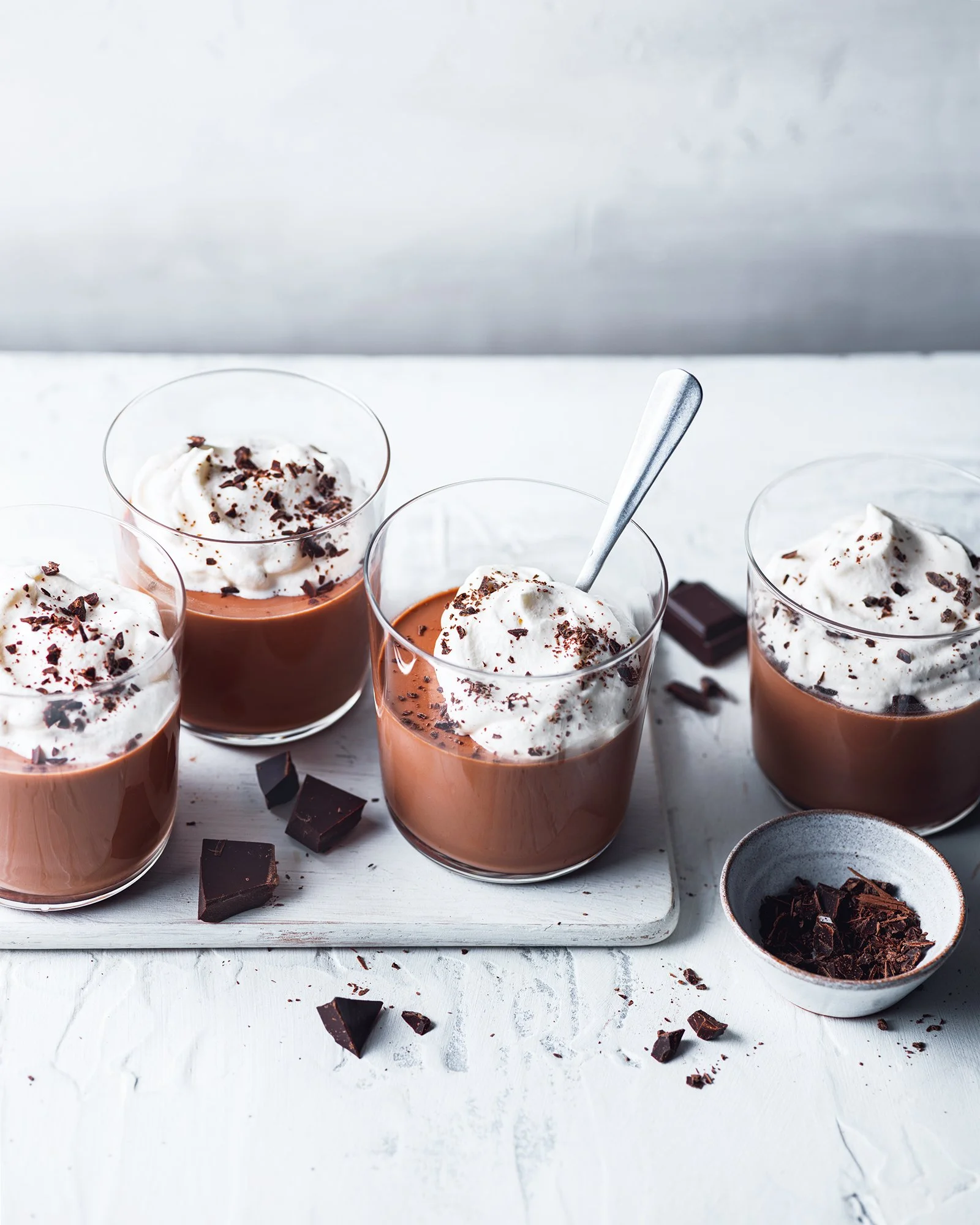 Four glasses of chocolate mousse topped with whipped cream and chocolate shavings on a white surface with chocolate pieces and a small bowl of chocolate shavings nearby.