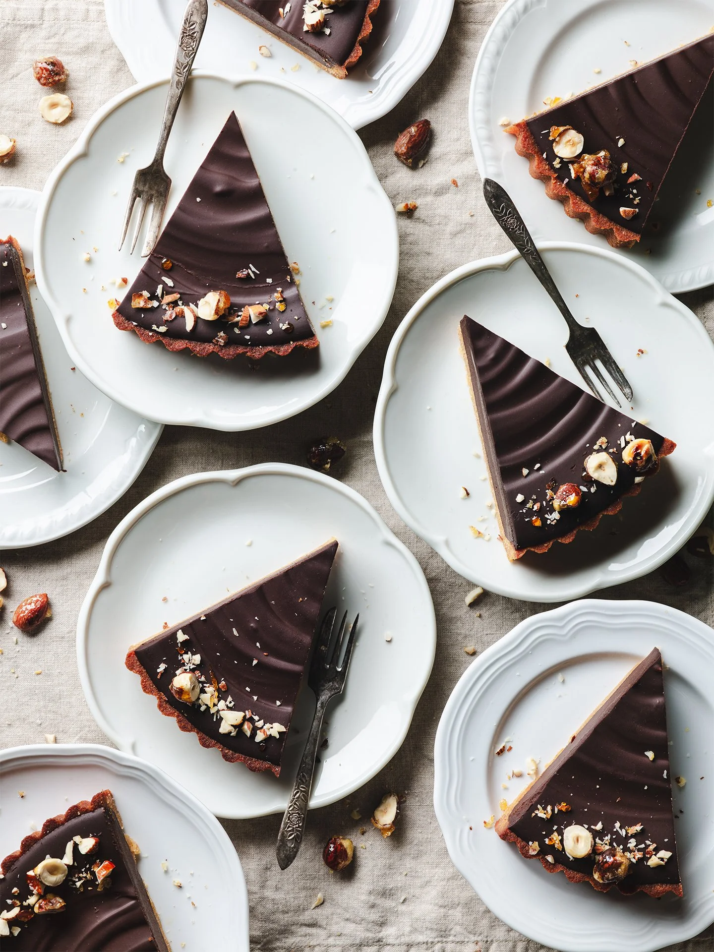 Slices of chocolate tart with nuts on white plates, set on a light fabric surface, some crumbs and scattered nuts around.