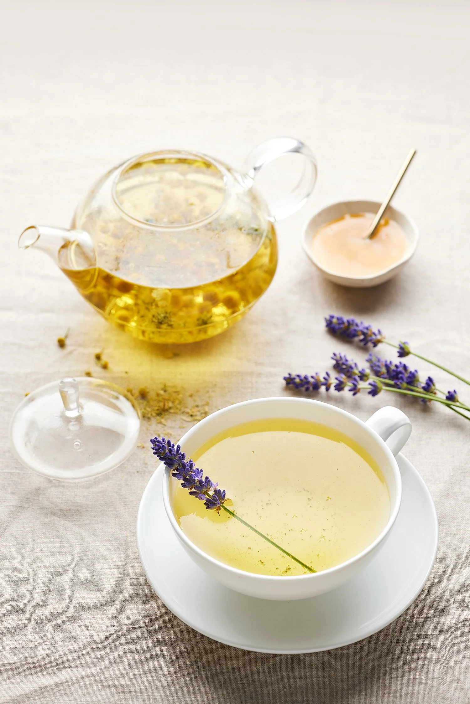 A white cup filled with lavender-colored tea, garnished with a sprig of lavender. A glass teapot with herbal tea and a small bowl with a spoon are nearby, with loose herbs scattered on a light-colored tablecloth.