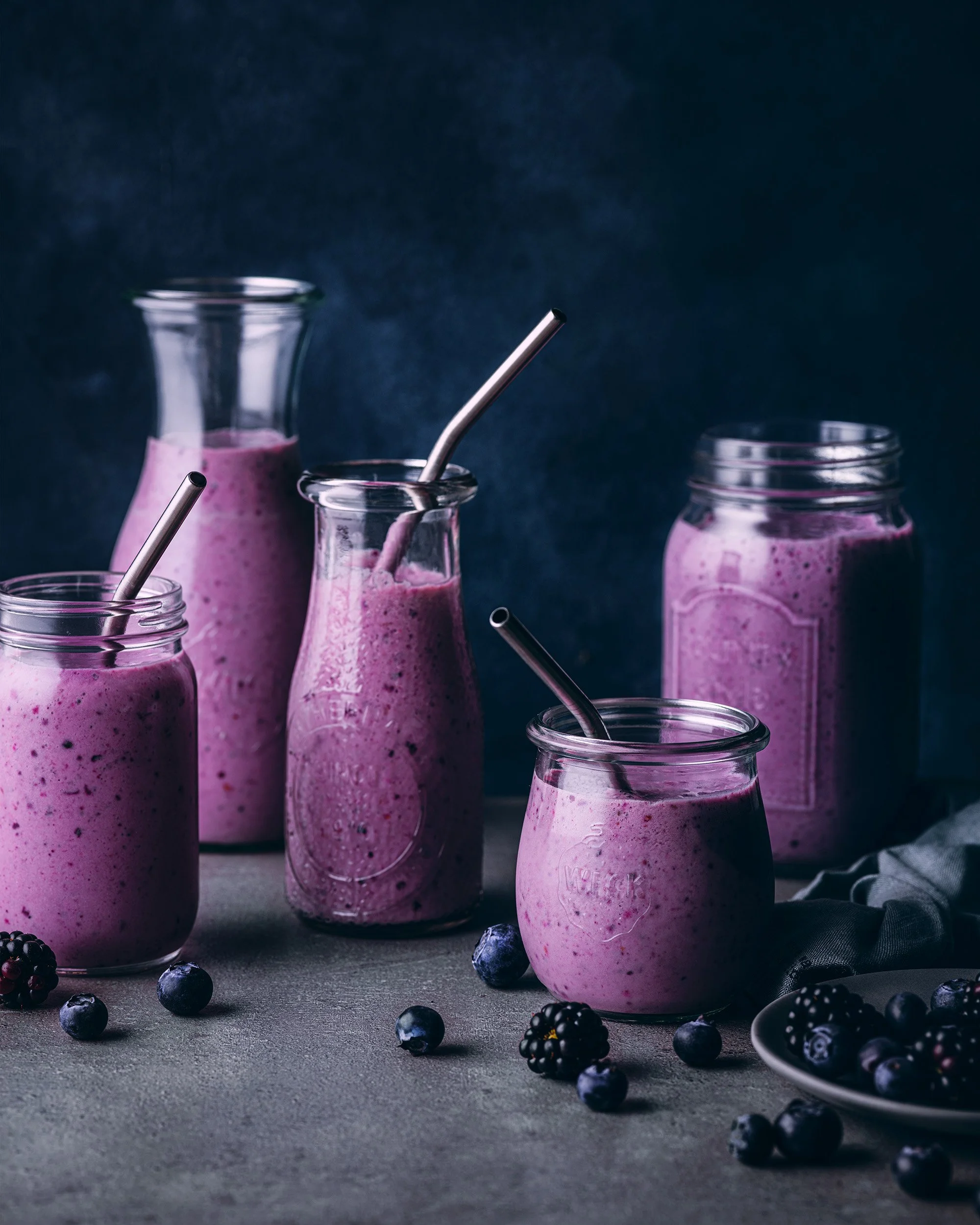 Several glass jars and bottles filled with pinkberry smoothies, with straws, surrounded by scattered blueberries and blackberries on a dark textured surface.