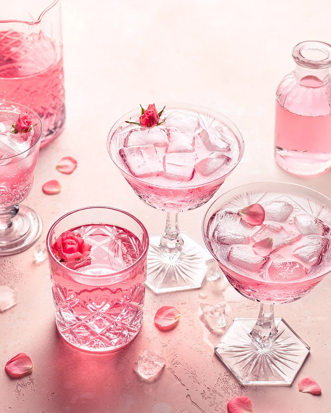 Four pink-colored drinks with ice cubes and pink rose petal garnishes in various glassware on a pink surface, with scattered rose petals and ice.