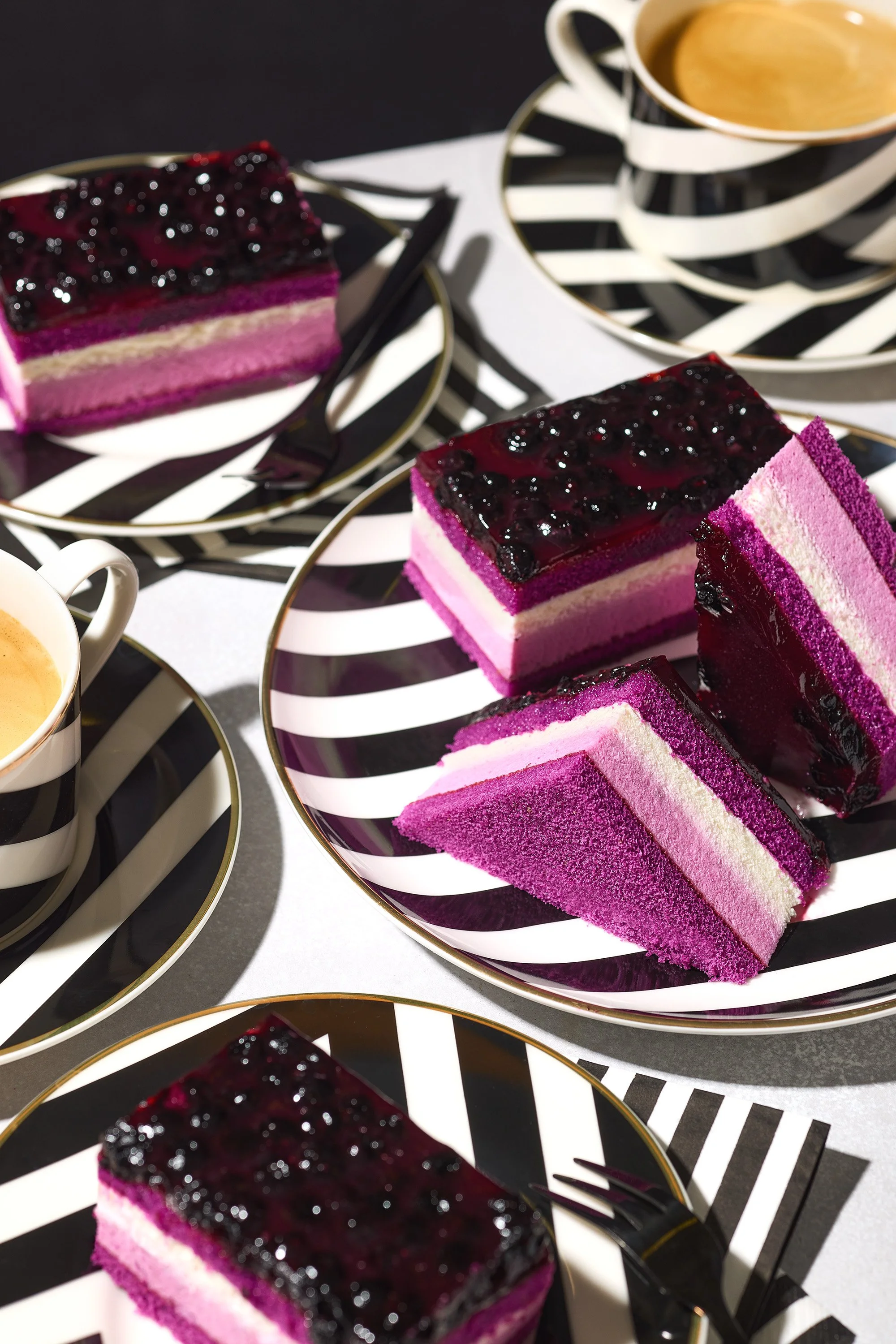 Slices of layered berry mousse cake on black and white striped plates with cups of coffee on matching saucers.