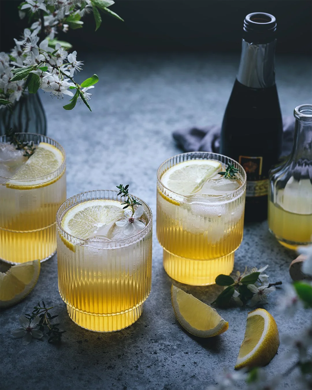 Three glasses of lemonade with lemon slices and flower garnishes on a gray surface, with lemons, a vase with white flowers, and bottles in the background.