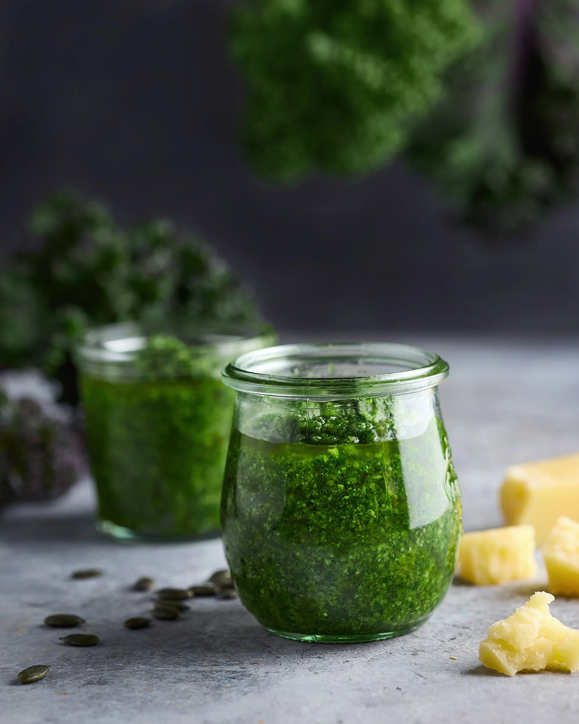 A close-up of a jar filled with green pesto, with chunks of cheese and pumpkin seeds on a gray surface in the foreground and more jars of pesto in the background.