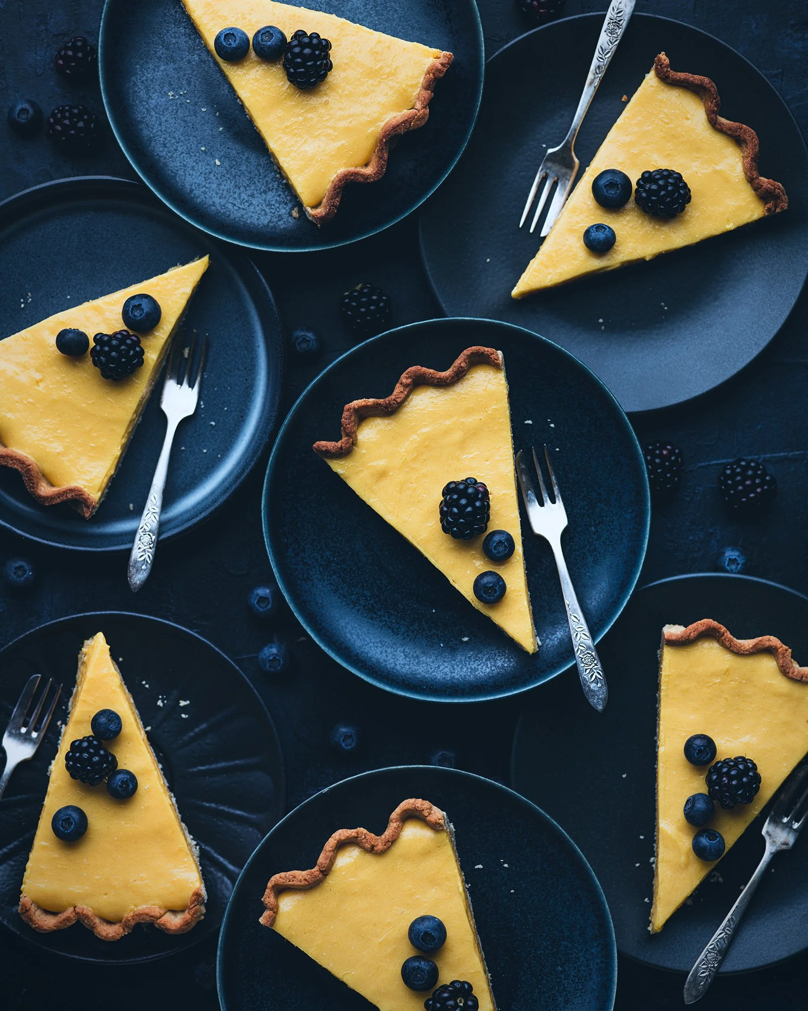 Several slices of lemon tart with blueberries and blackberries on dark plates, with small forks on each plate, arranged on a dark surface.