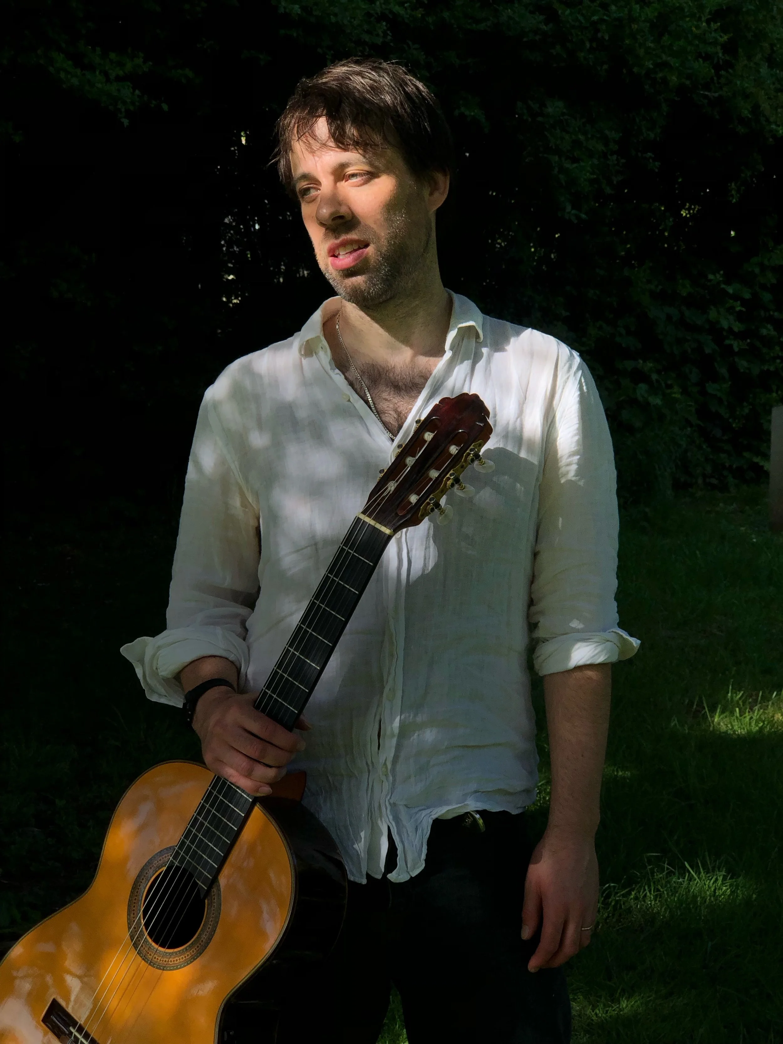 Nicolas Lewis classical guitarist holding guitar outside in dappled shade before performing in York
