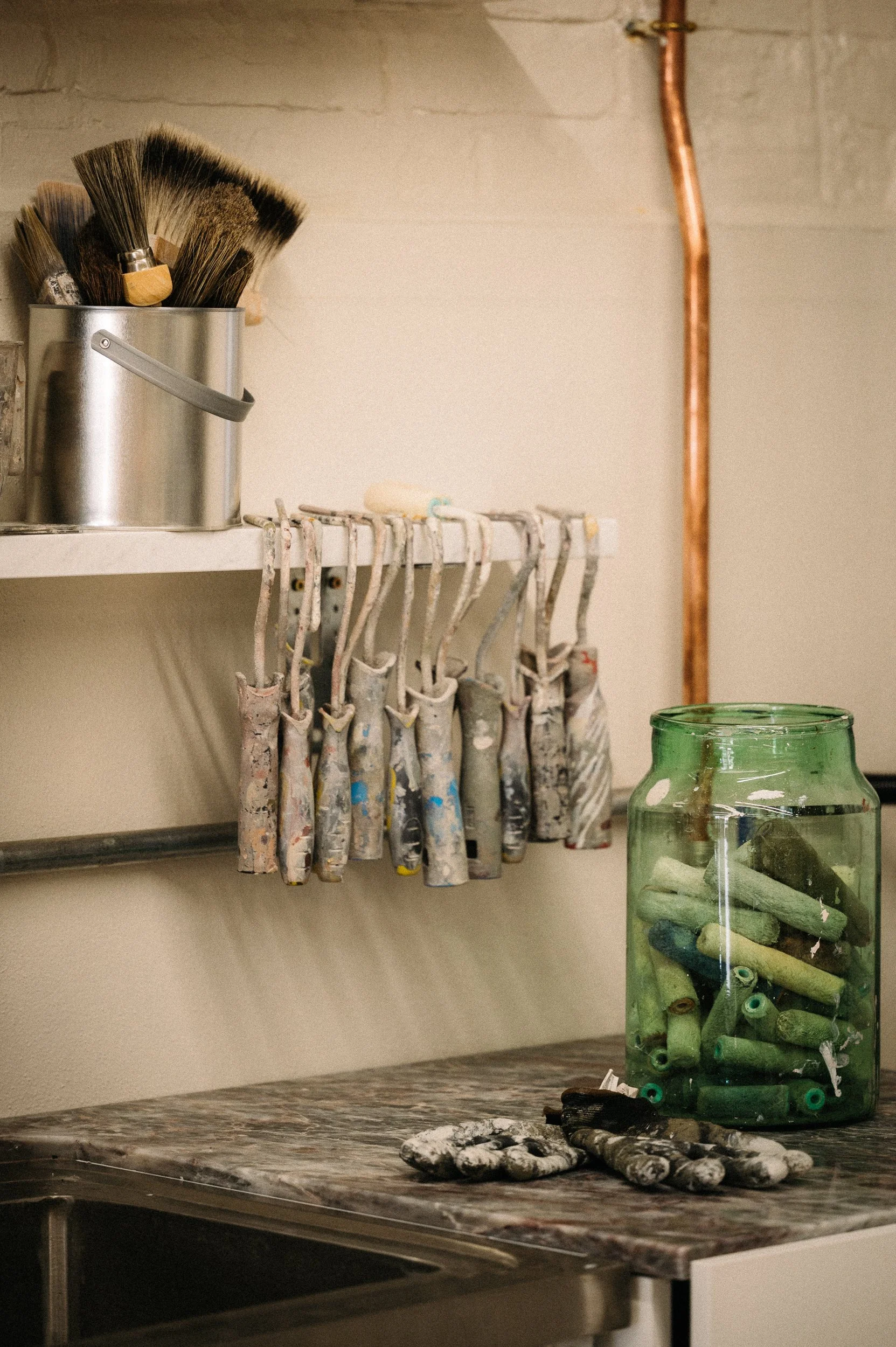 Atelier Ellis Studio - Paintbrushes in a metal container on a shelf, paint rollers and gloves on a marble countertop, and a jar filled with used chalks.