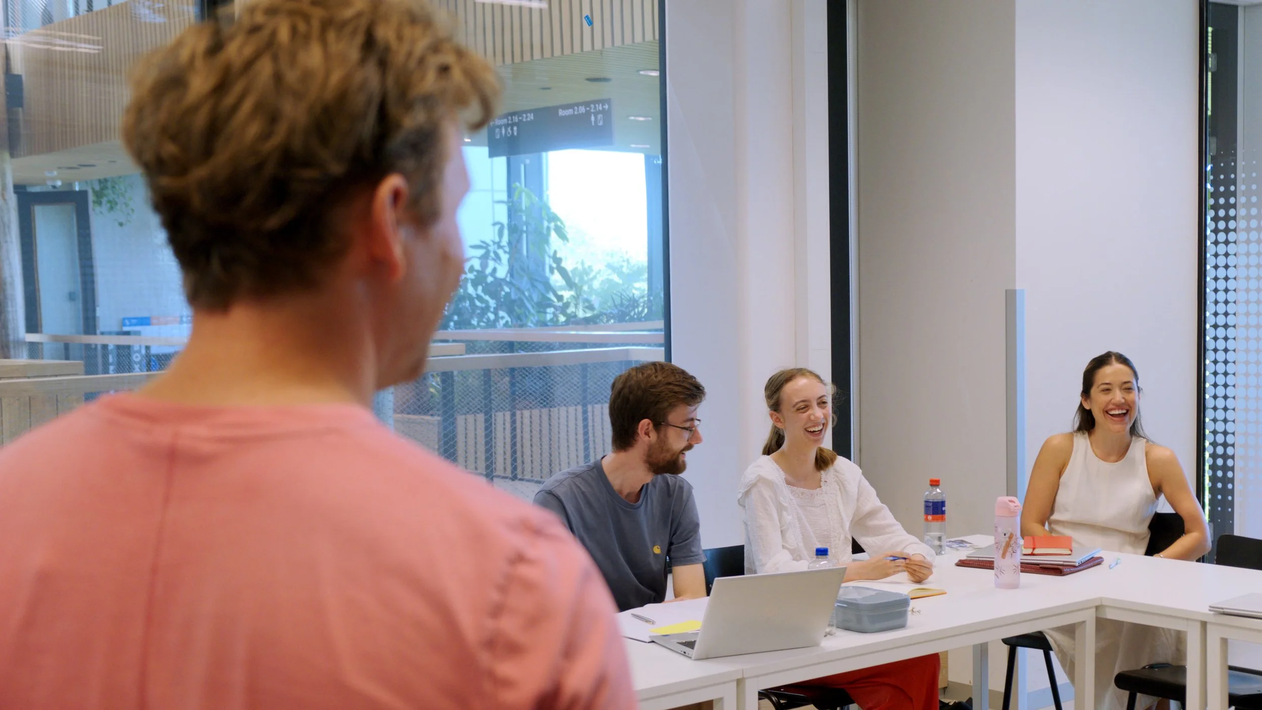 A group of people laughing and talking together during a meeting in a modern office, with laptops, notebooks, and water bottles on the table.