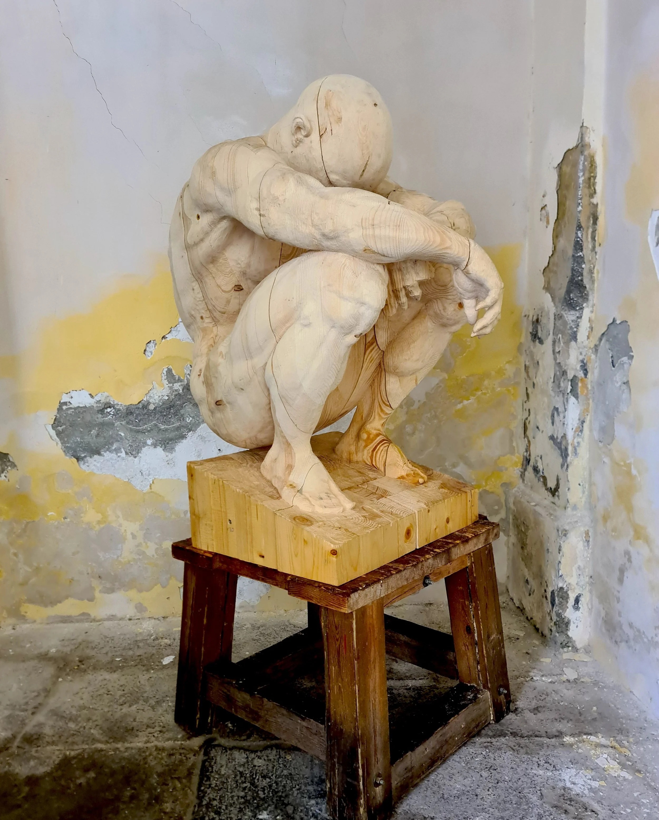 Statua di legno di un uomo seduto con le braccia sulle ginocchia, posizionata su un supporto in legno, in un ambiente con pareti usurate e con alcune crepe.