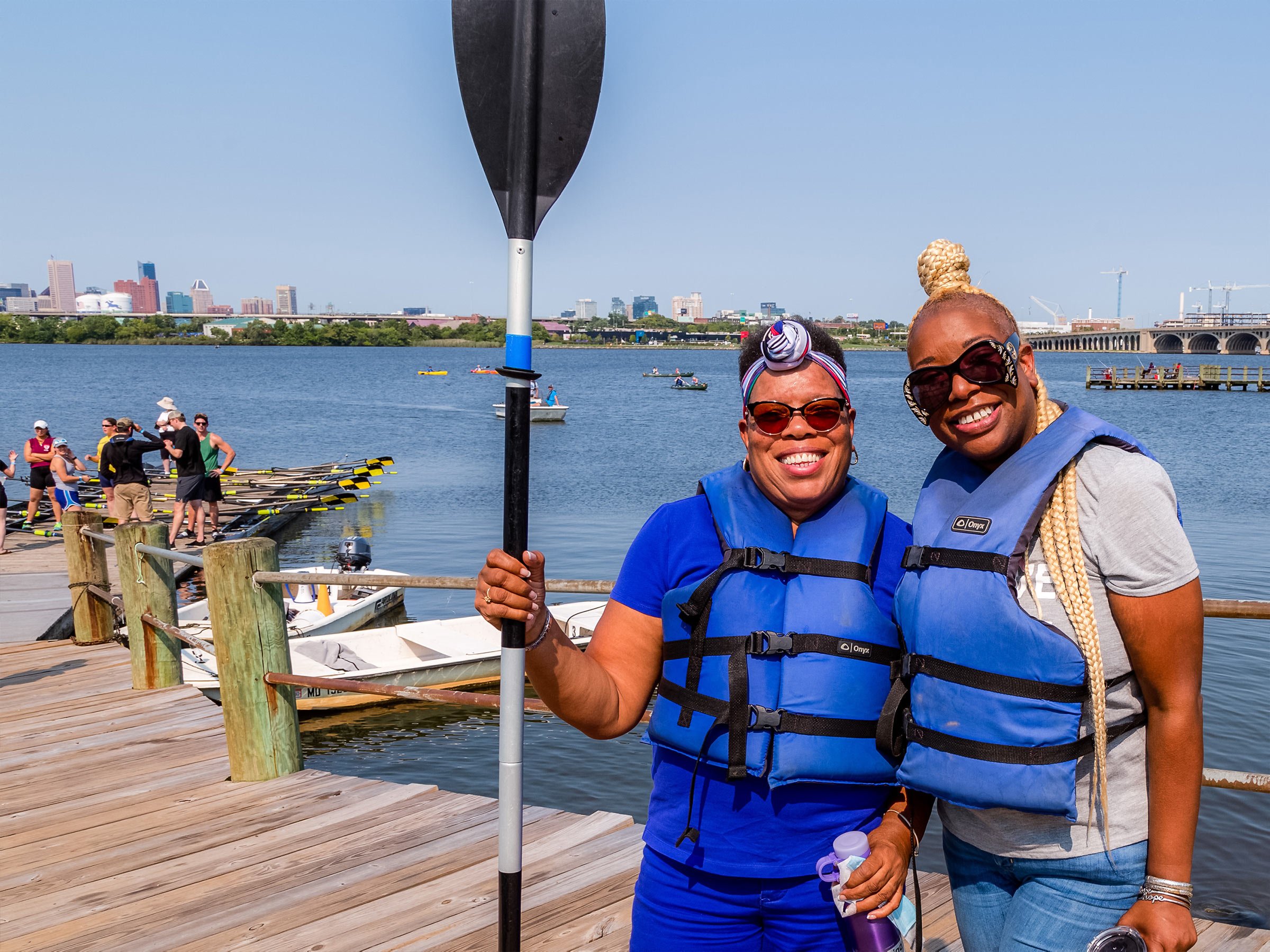  Two women smile together on boathouse dock. They hold a paddle and sport life vests. People in canoes and kayaks are behind them. 