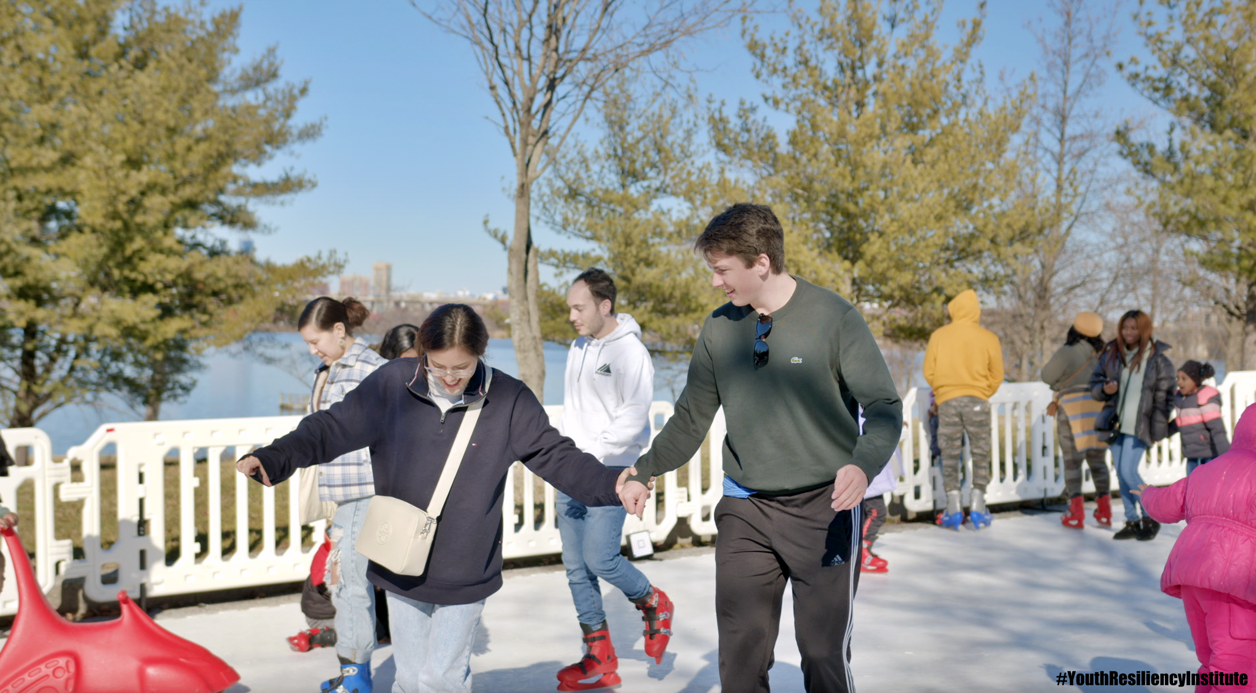  People are ice-skating and holding hands on the ice-rink with a view of the Middle Branch waterfront in the background. 
