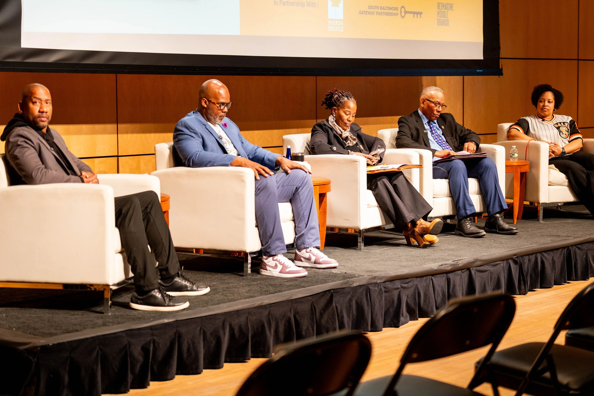  Five panelists sit in white chairs on stage during the event.  