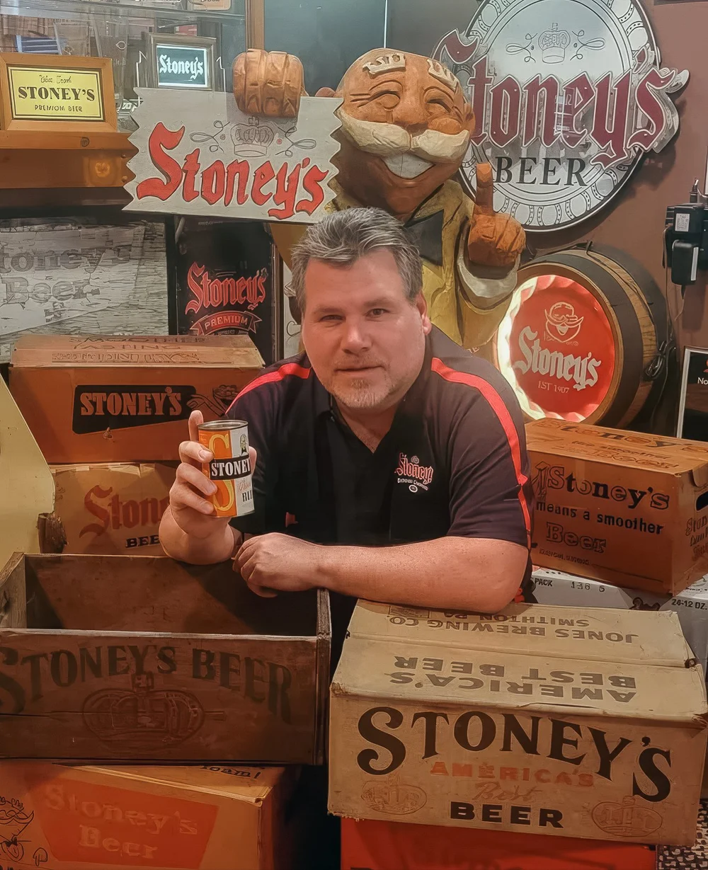 Bill Stoney holding a can of Stoney's Beer in his collection room, surrounded by vintage signs and wooden crates