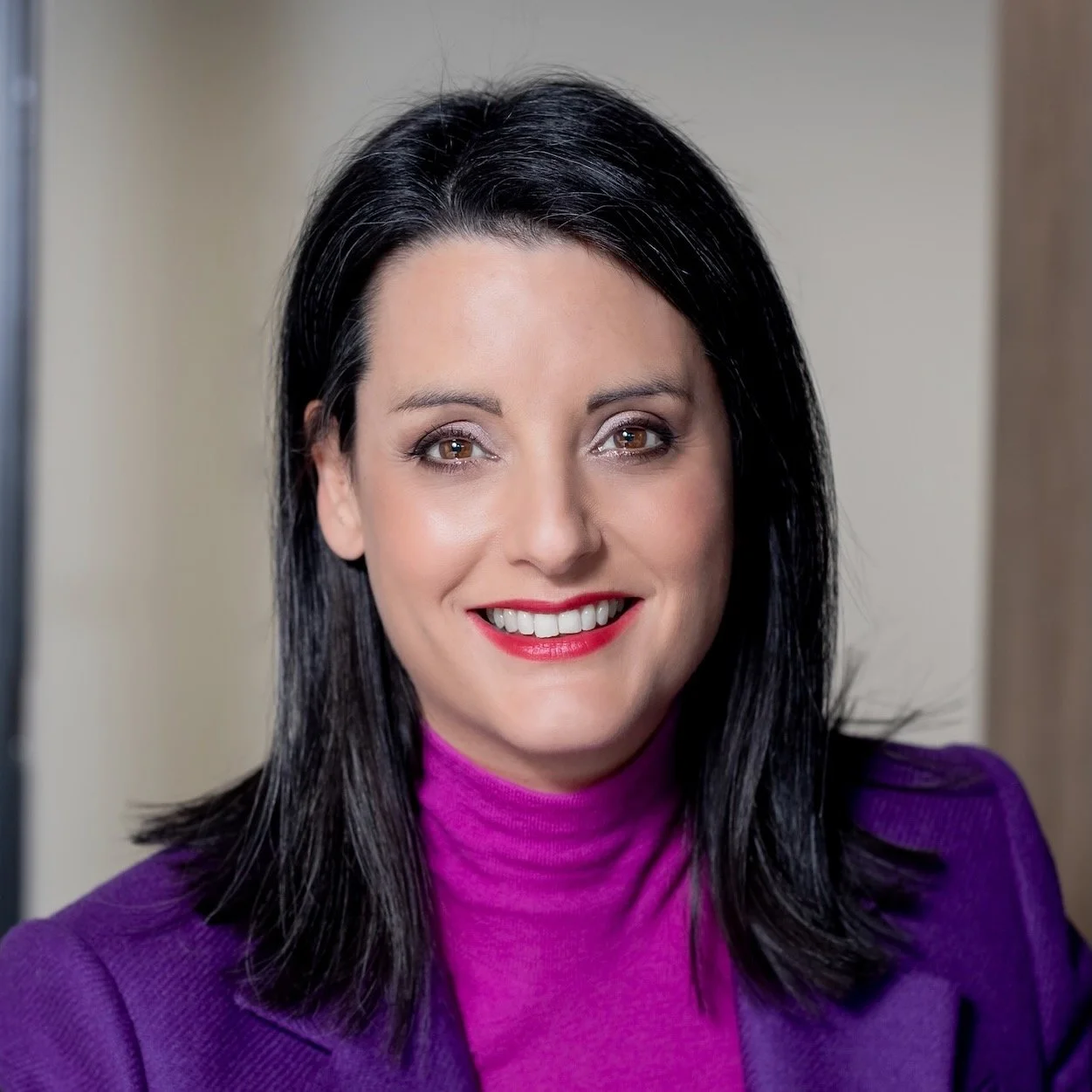 A woman with black hair and brown eyes smiling, wearing a purple top with matching lipstick, in an indoor setting.