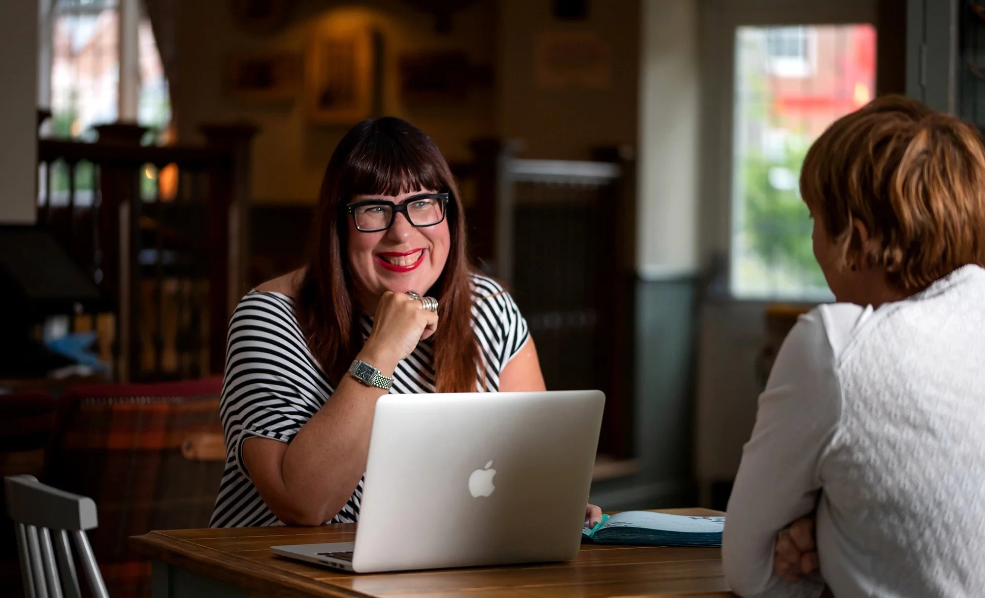 Two women sitting at a wooden table in a cozy cafe, smiling and engaging in conversation, with a laptop and open book on the table.